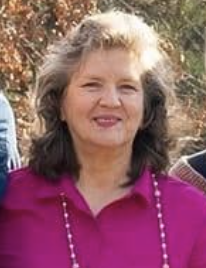 A woman with shoulder-length gray hair, wearing a bright pink top and a pearl necklace, standing outdoors with trees in the background.