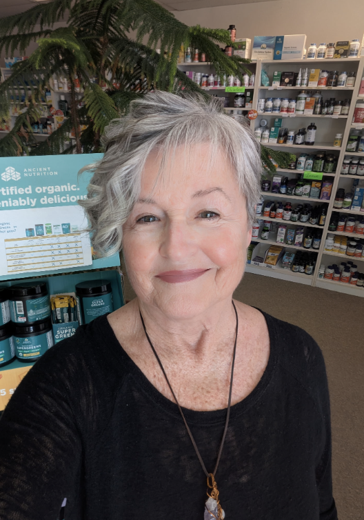 Smiling elderly woman with short gray hair taking a selfie inside a health supplement store. Shelves filled with bottles and containers of vitamins and supplements are visible in the background.