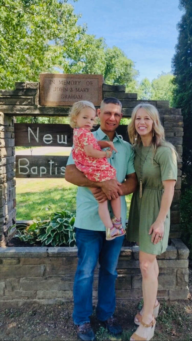 A family of three standing outdoors in front of a sign that reads "New Baptist," with a memorial plaque above and trees in the background. The man is holding a young girl in a pink dress, and a woman in a green dress is standing beside them, smiling.