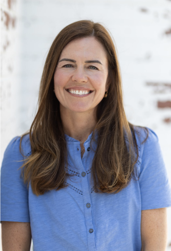 A woman with long brown hair smiling, wearing a blue button-up shirt, standing in front of a white brick wall.