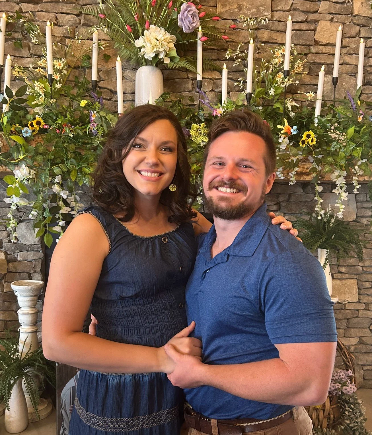 A smiling woman and man holding each other in front of a decorative floral arrangement and candle holders against a stone wall.