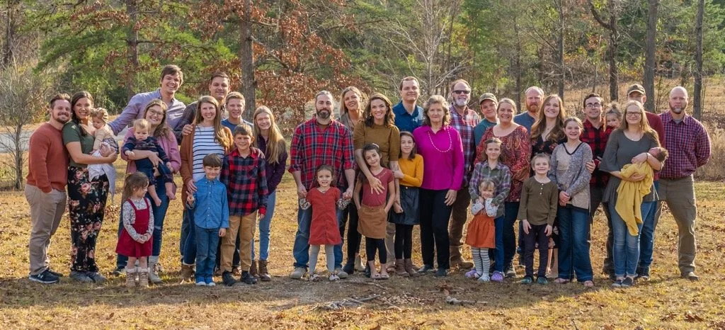A large family gathers outdoors in a park during fall, standing on grass with trees in the background, posing for a group photo.