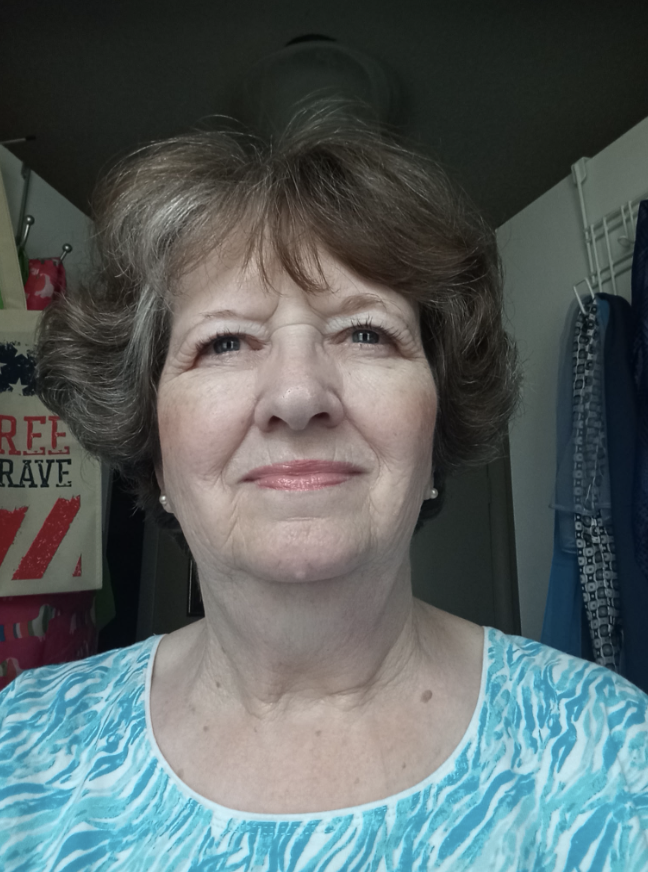 A middle-aged woman with short, curly hair taking a selfie indoors. She is wearing a blue and white patterned top and has a slight smile. In the background, there are clothing and accessories hanging, and a ceiling with a vent.