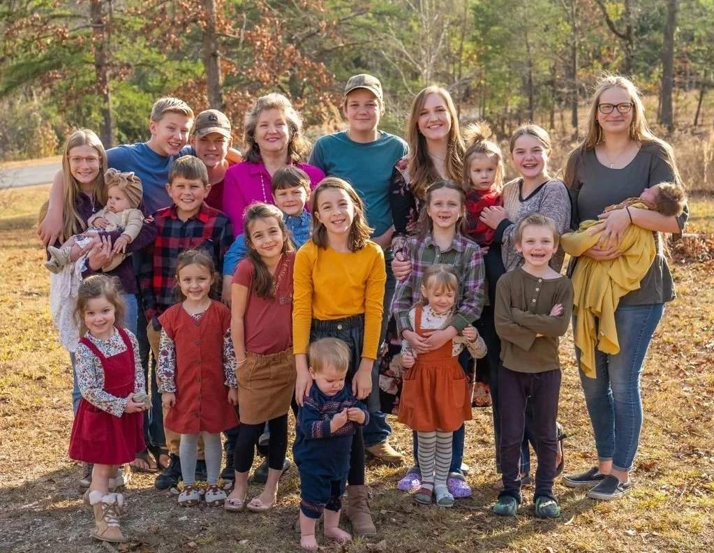 A large family group posing outdoors during autumn, with fall leaves in the background. The group includes children and adults, some holding babies, all smiling.