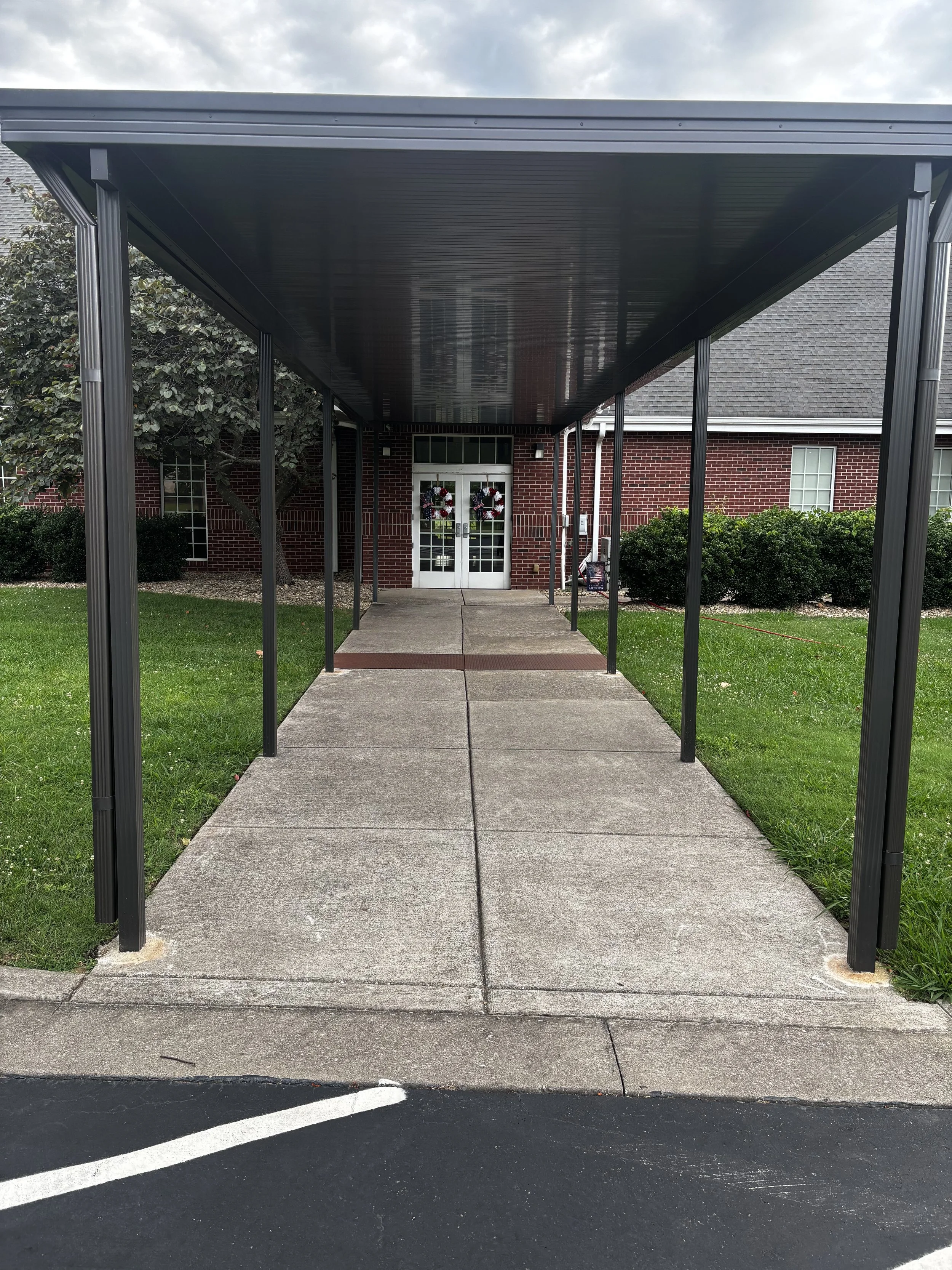 Sidewalk walkway through a black metal covered pathway leading to a brick building with front doors decorated with holiday wreaths, surrounded by neatly trimmed bushes and grass, with cloudy sky overhead.