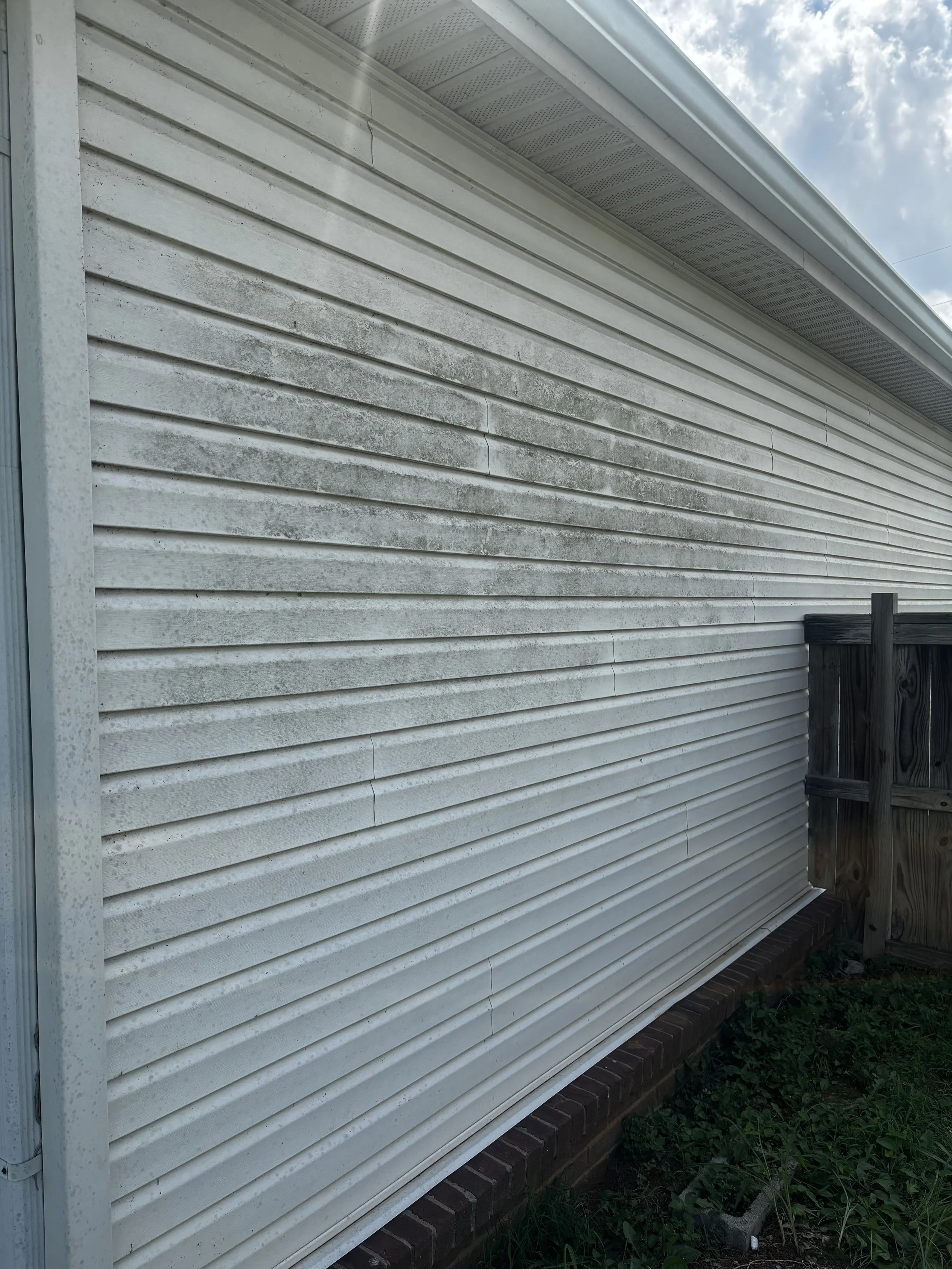 The exterior of a house with white vinyl siding, a brick foundation, and a wooden fence on the right side. The sky is partly cloudy.