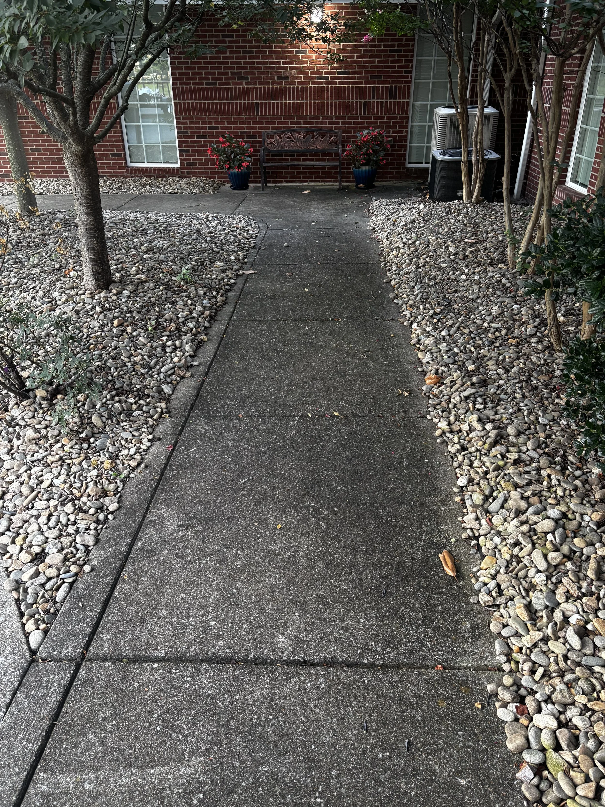 Concrete sidewalk leading to a brick house with a gate in the background. Flanked by landscaping with small rounded stones, trees, and bushes. Two flower pots with pink flowers and a bench are positioned against the house.