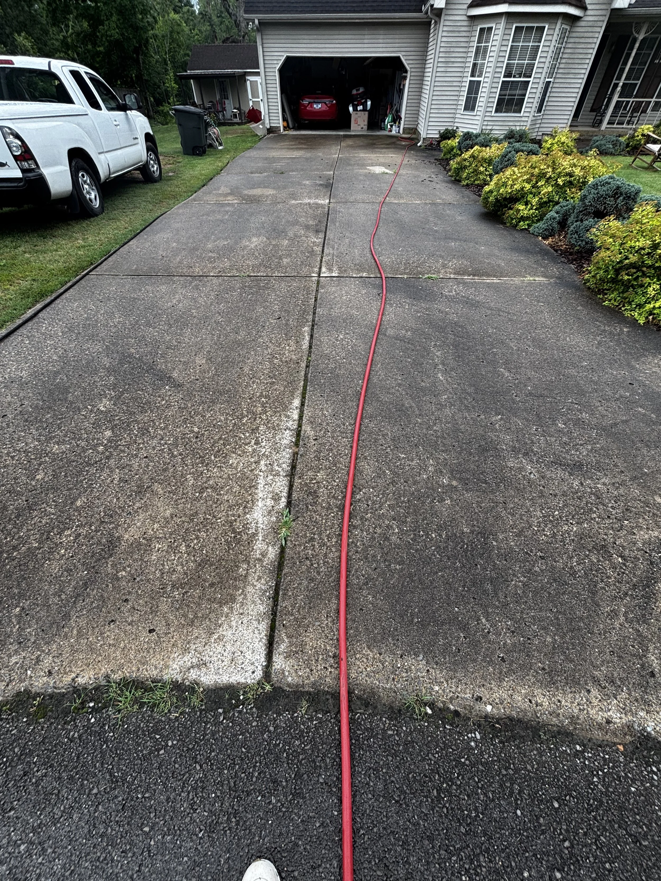 View of a concrete driveway leading to an open garage attached to a house with gray siding. A red hose runs along the driveway, and a white pickup truck is parked on the grassy area to the left. There are various items, including a trash bin and bicy