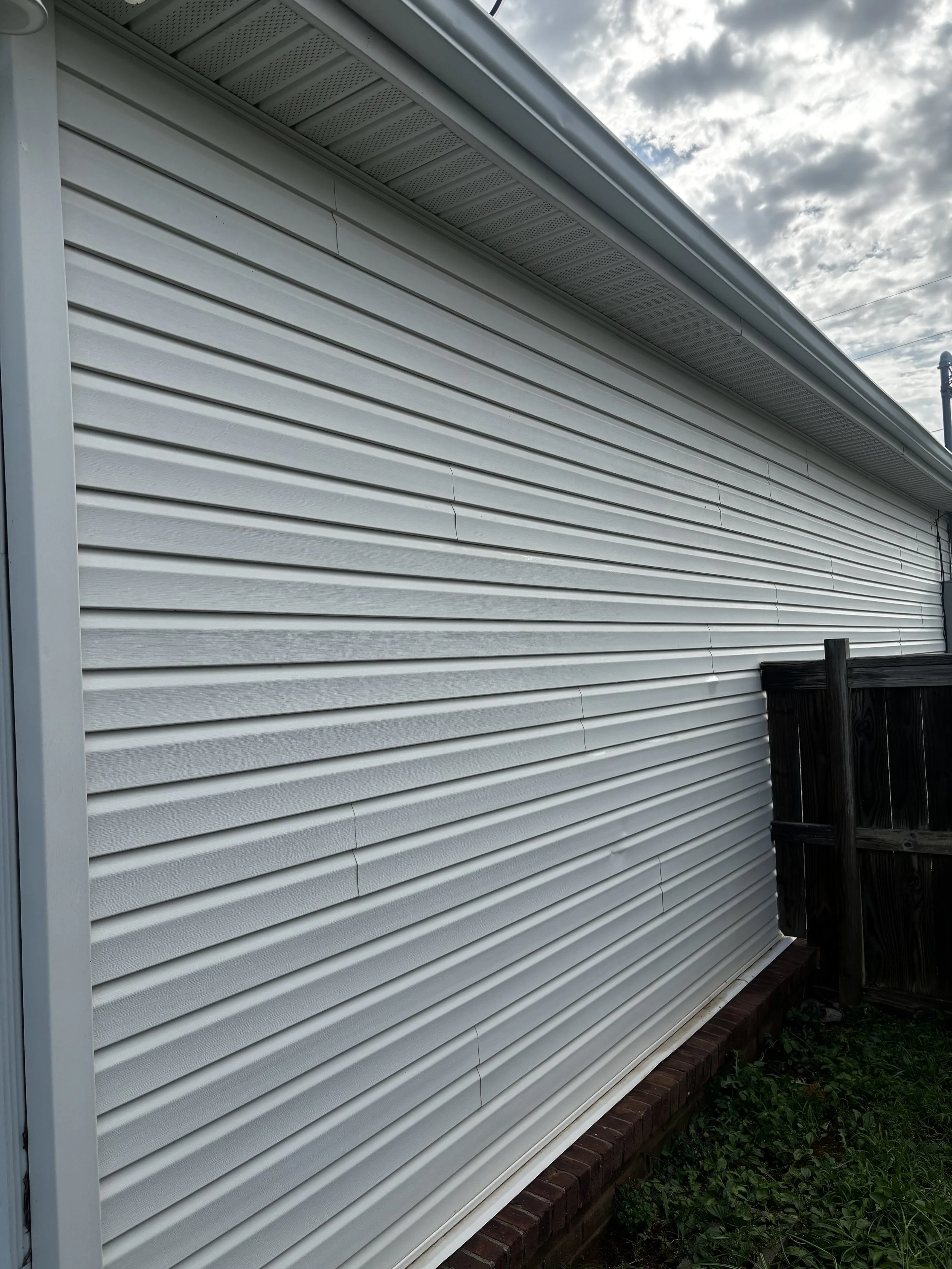 Side view of a house with white vinyl siding and a brick foundation, with a wooden fence and green plants at the bottom, under a cloudy sky.