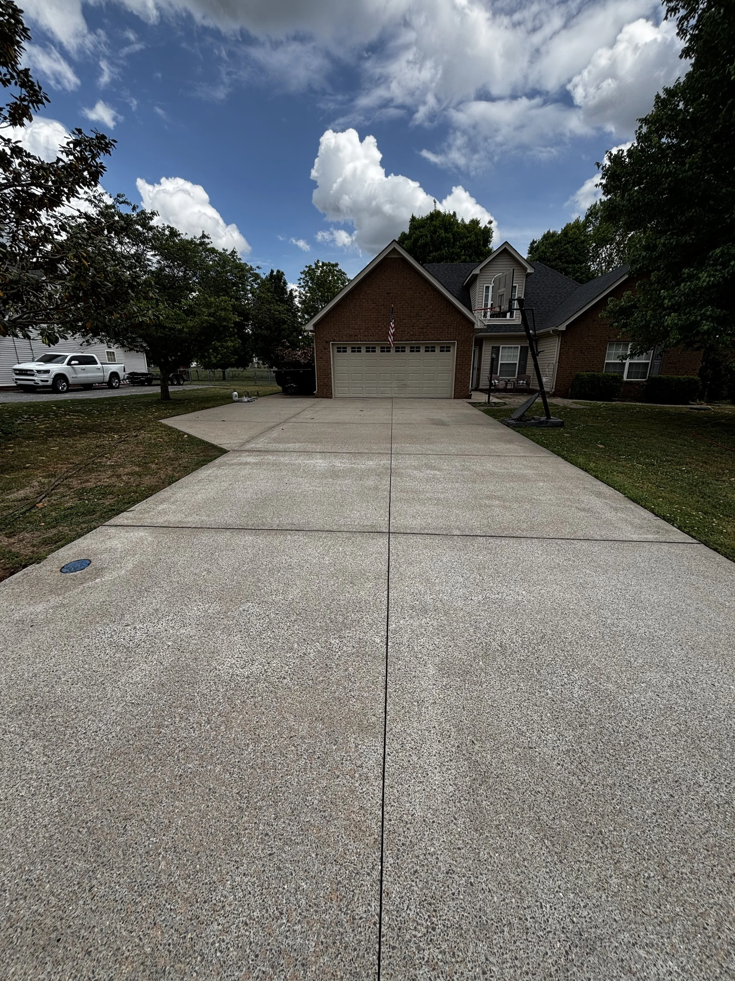 A residential driveway with a house in the background, a basketball hoop, a soccer ball, and two chairs near the house. The sky is partly cloudy.