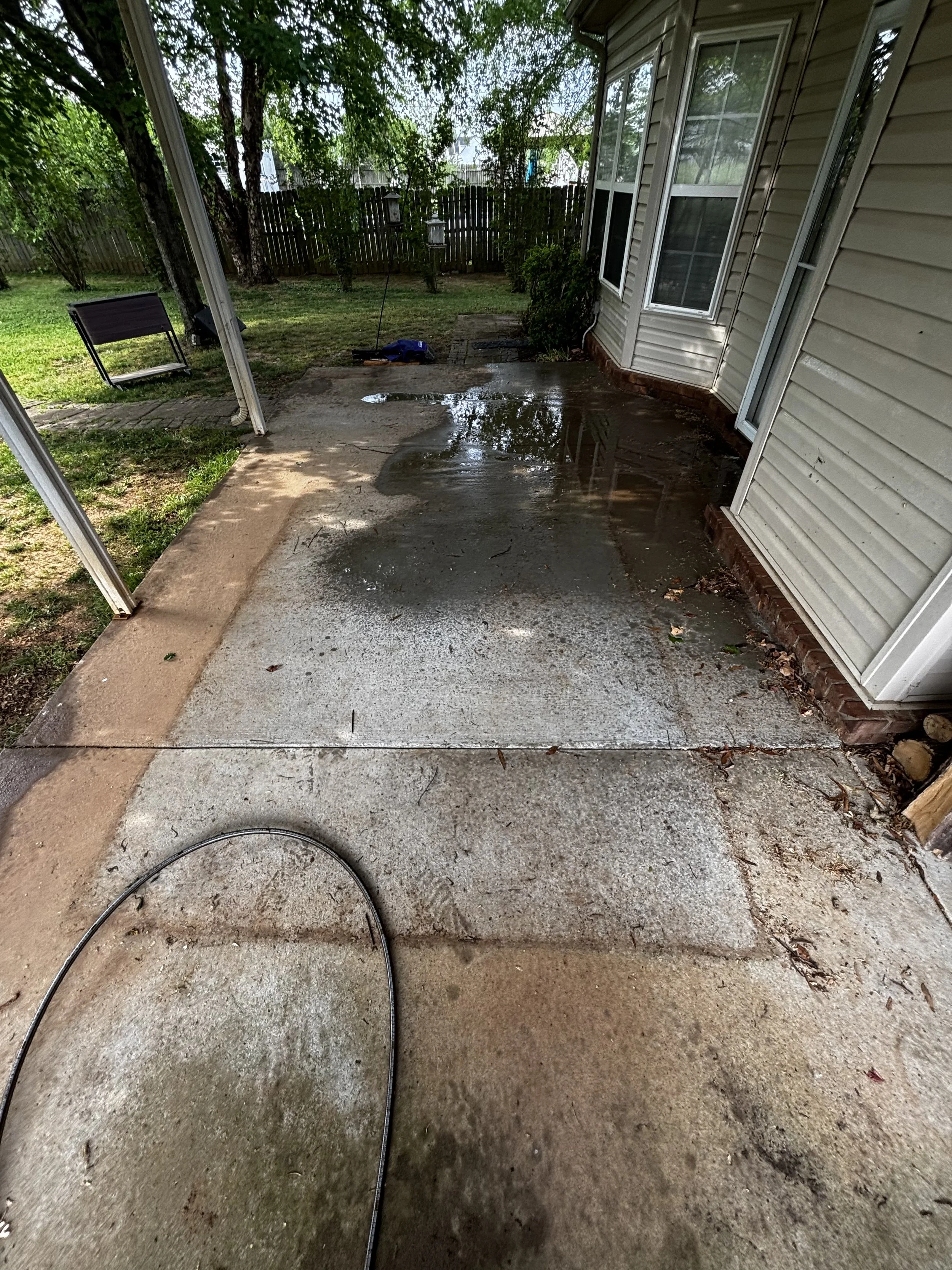 Wet concrete patio area outside a house with a white siding exterior, windows, and a sliding glass door. There is a black hose on the ground, some leaves and dirt, and water puddles on the surface. The yard has grass, trees, and a wooden fence.