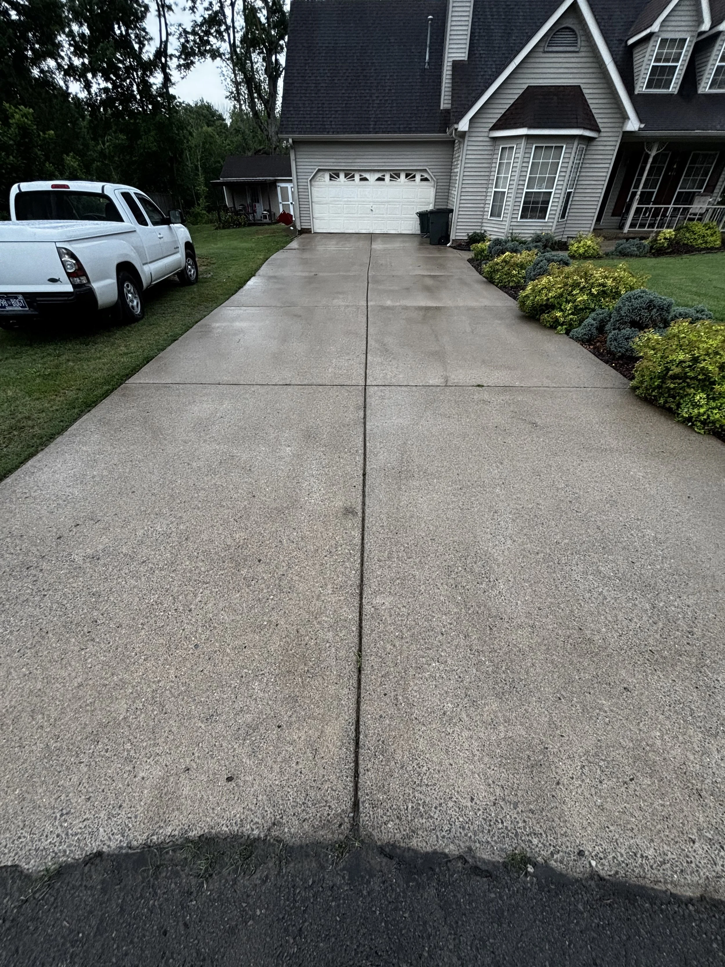 A wet concrete driveway leading to a house with a garage, with a parked white truck on the left side and landscaped bushes on the right side.