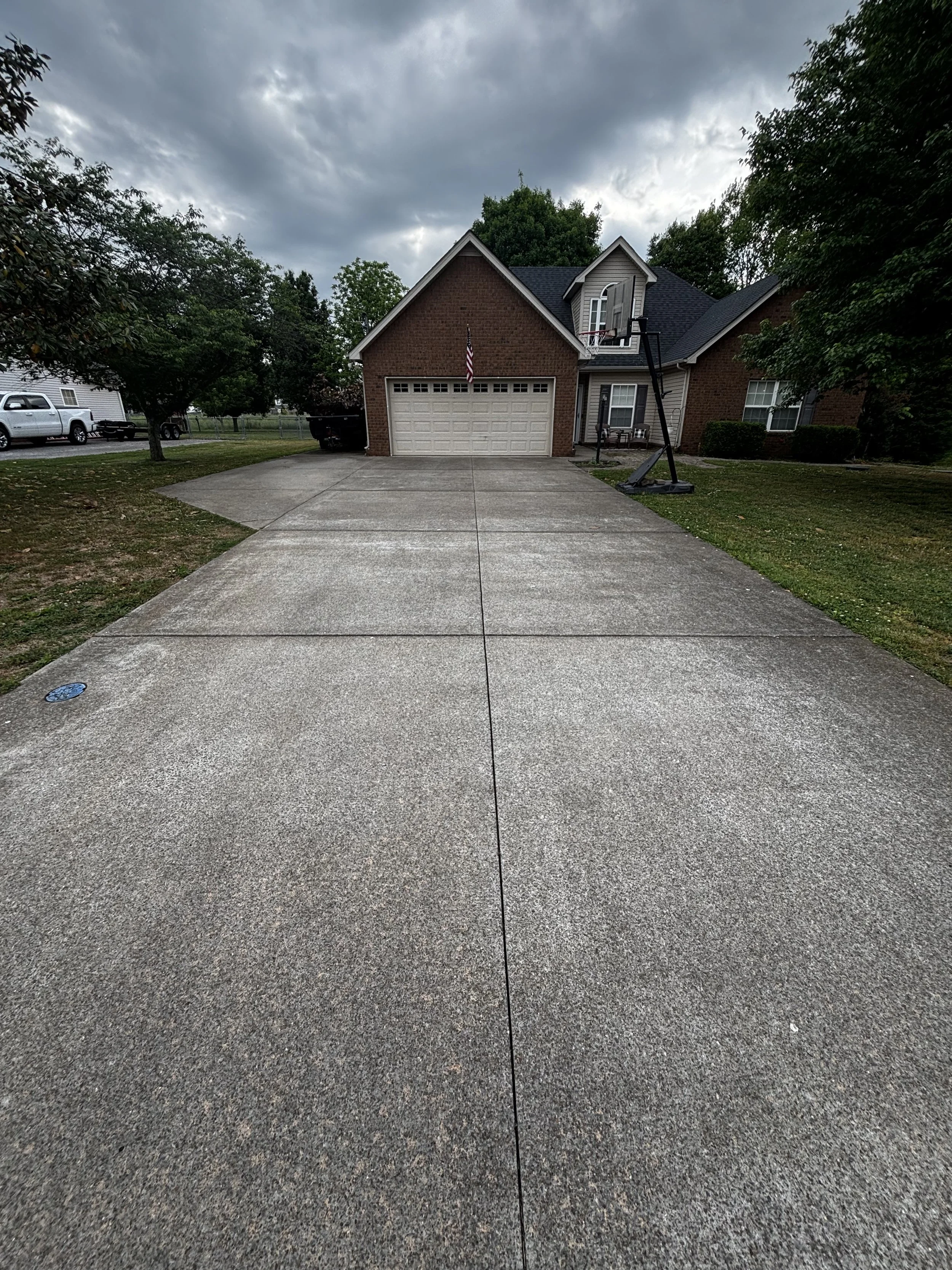 View of a residential house with a driveway, brick and siding exterior, and a basketball hoop on a tall stand in the driveway, under a cloudy sky with trees and parked vehicles nearby.