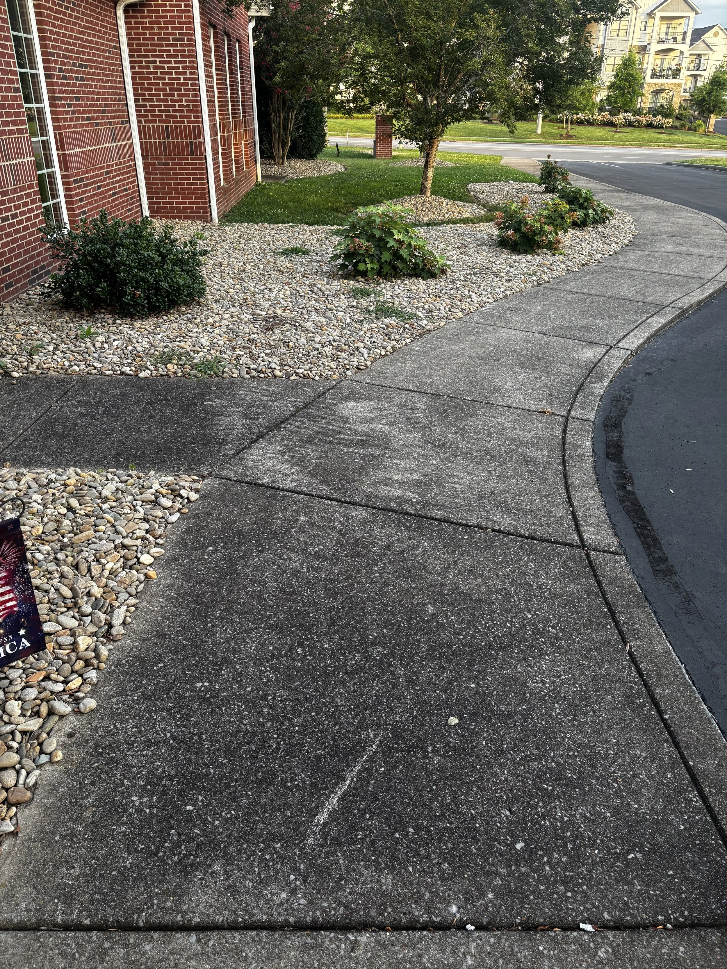 A sidewalk bordered by a landscaped area with small rocks, bushes, and a tree, in front of a brick building