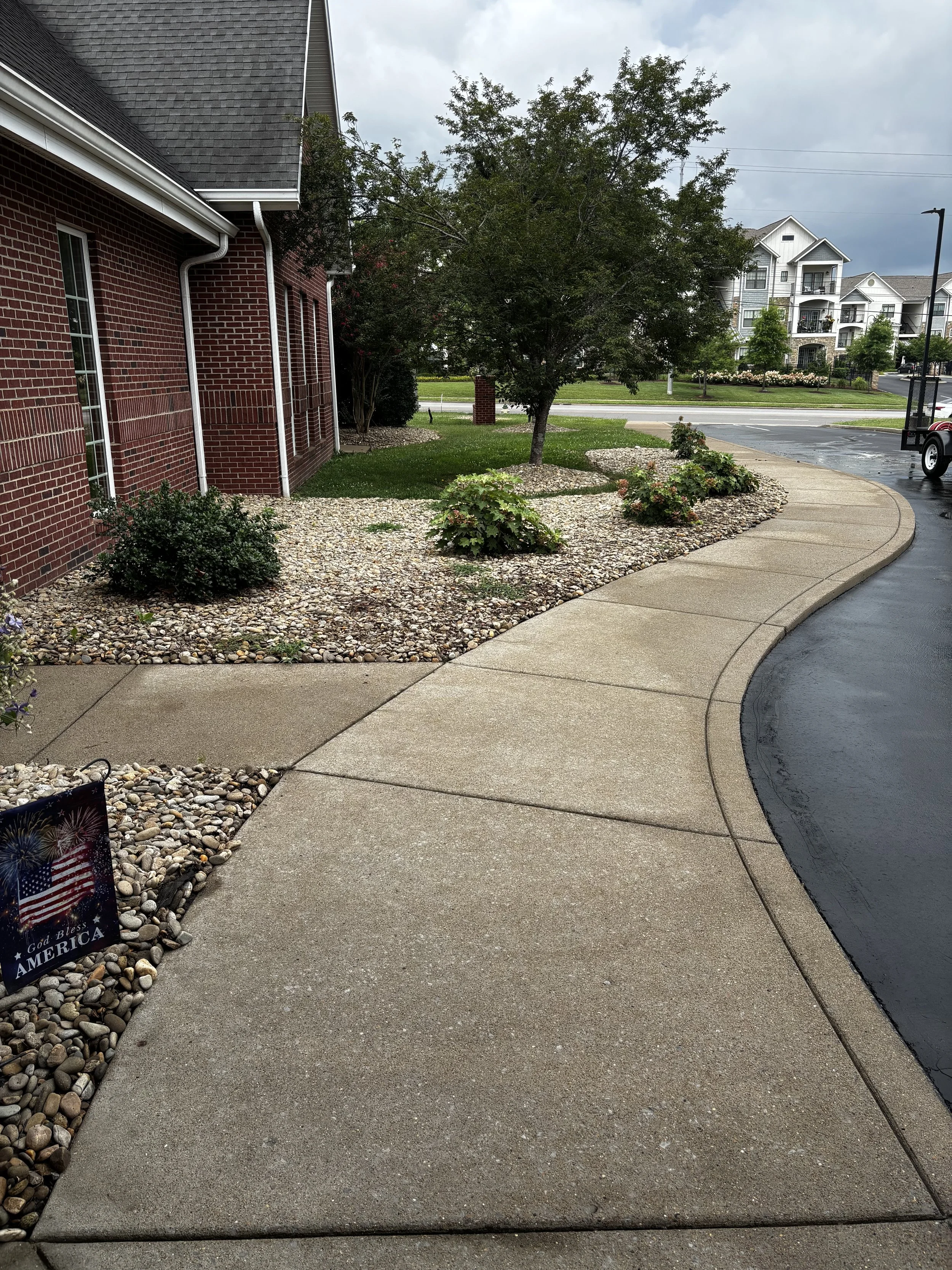 A sidewalk next to a brick building with a small garden area containing shrubs and a tree, with a parking lot and residential buildings in the background under cloudy skies.