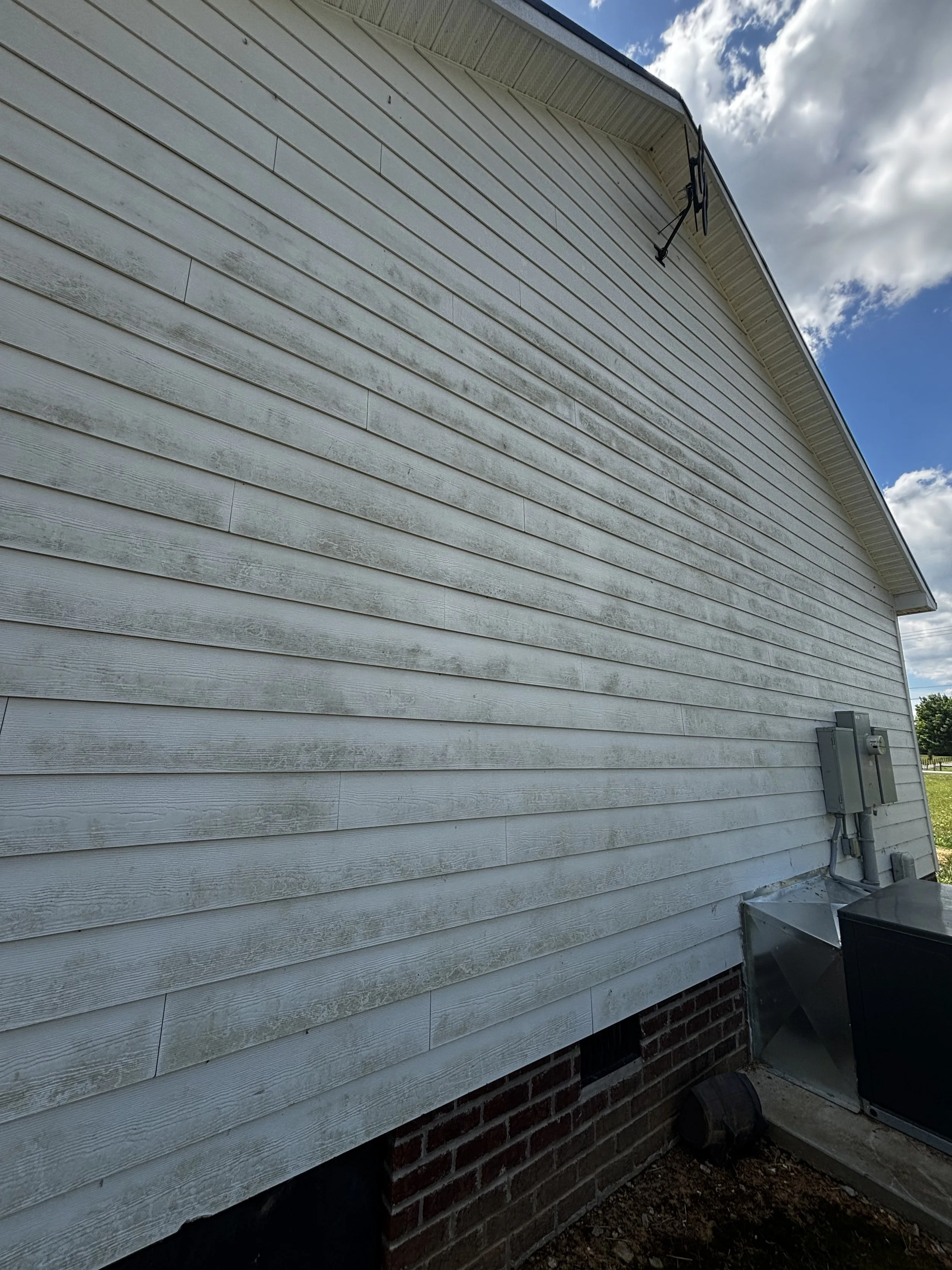 Exterior view of a house with weathered light gray siding, gutter system, and electrical box, with a partly cloudy blue sky.