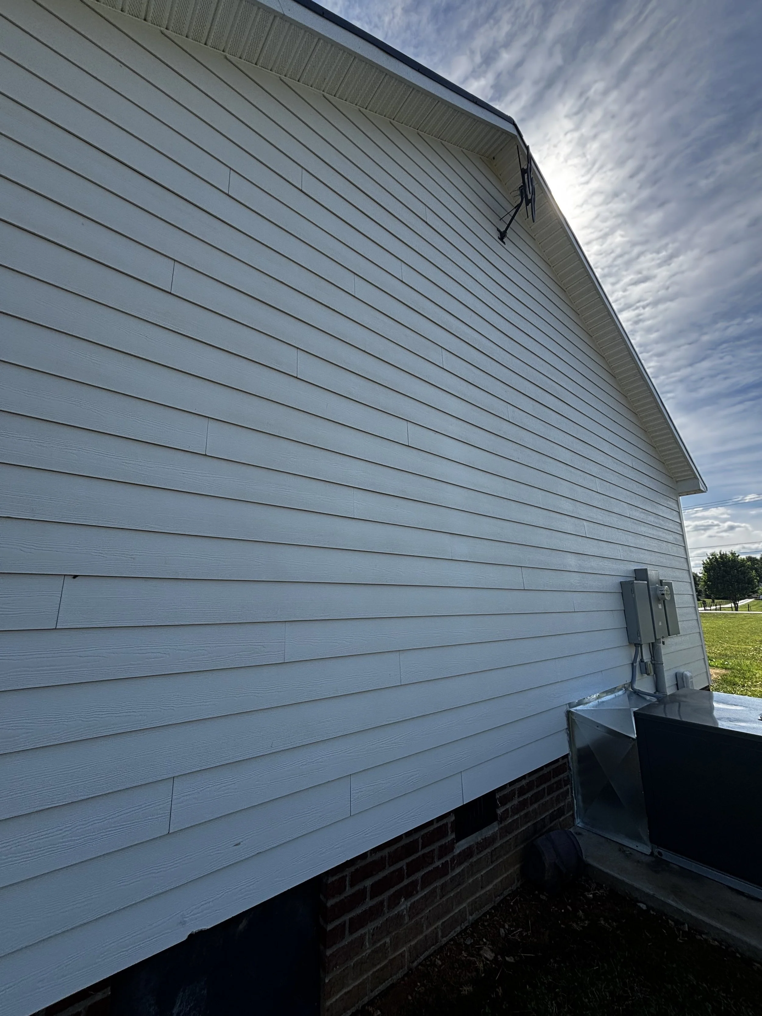 Side of a house with white horizontal siding, electrical meters, and an HVAC unit in the backyard, under a cloudy sky.