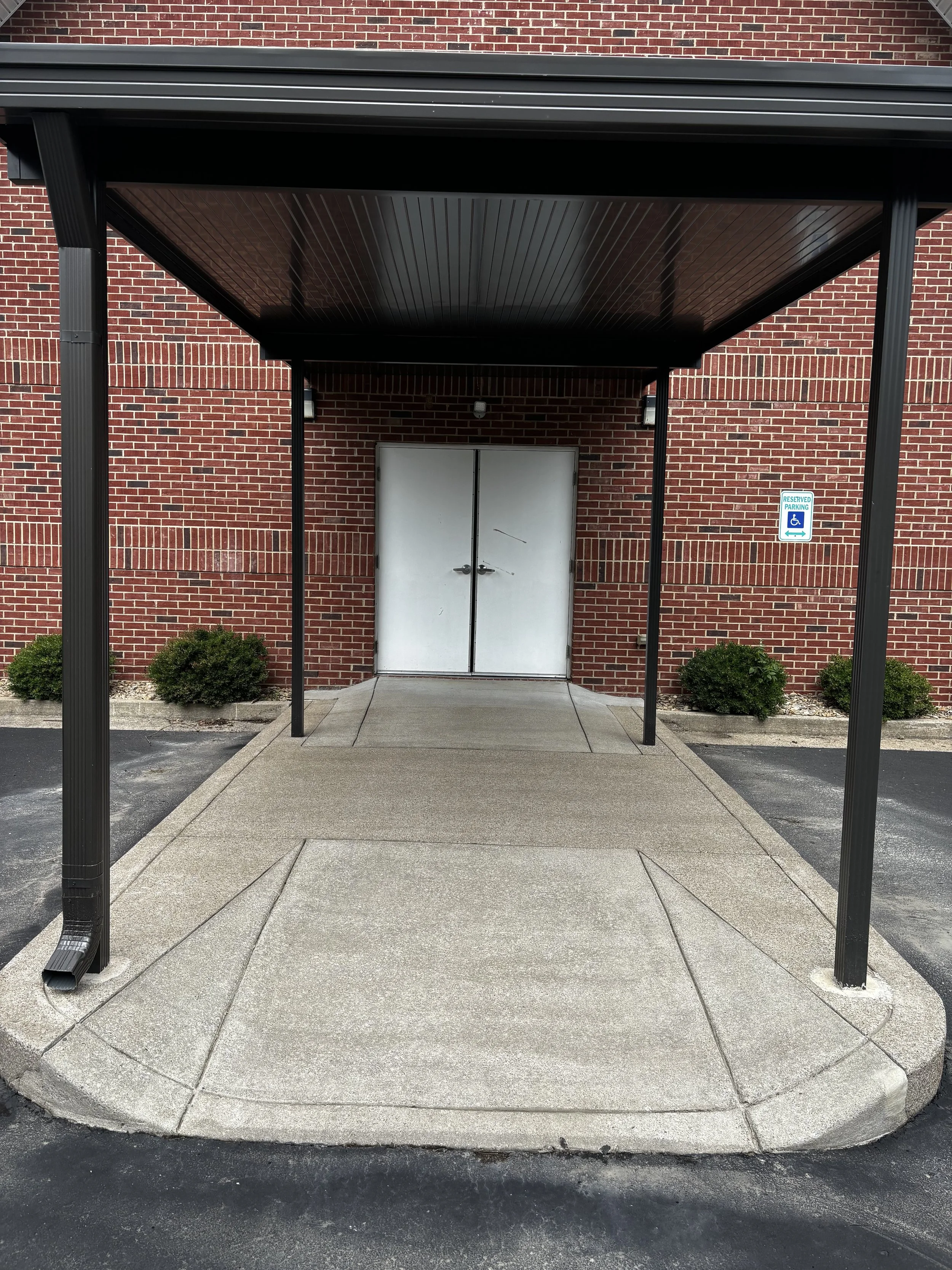 A building entrance with a metal covered walkway, two black metal posts supporting the roof, a double metal door, and a small reserved parking sign for handicapped parking on the brick wall to the right.