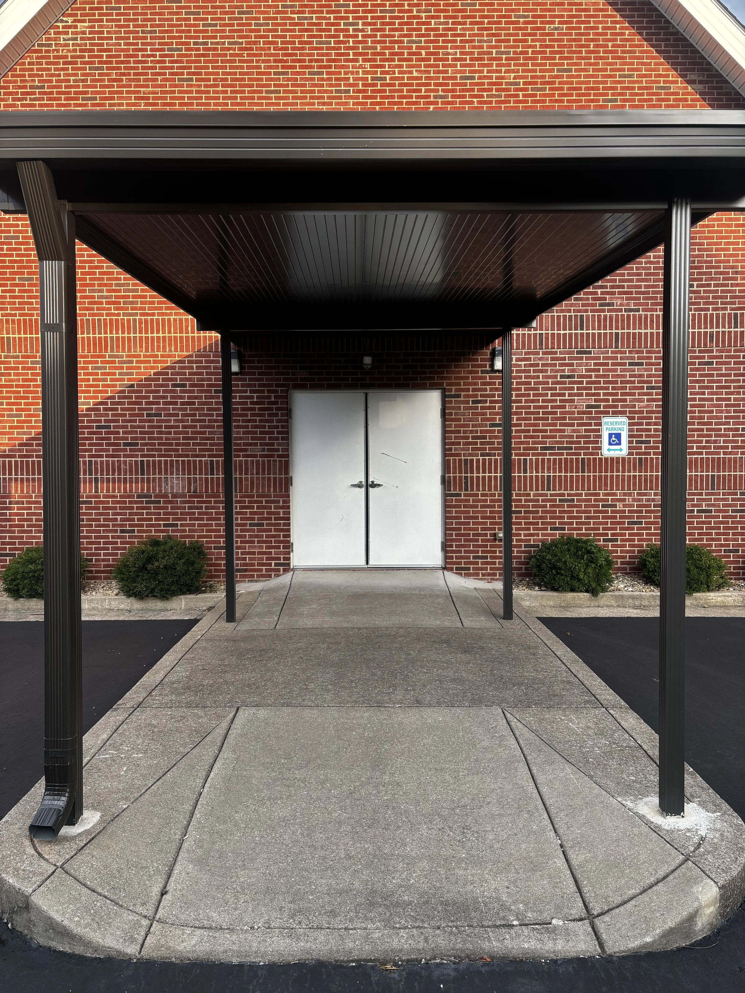 A brick building with a metal awning over a wheelchair-accessible entrance, marked by a blue and white sign, with shrubbery on each side of the door and concrete sidewalk and curb in the foreground.