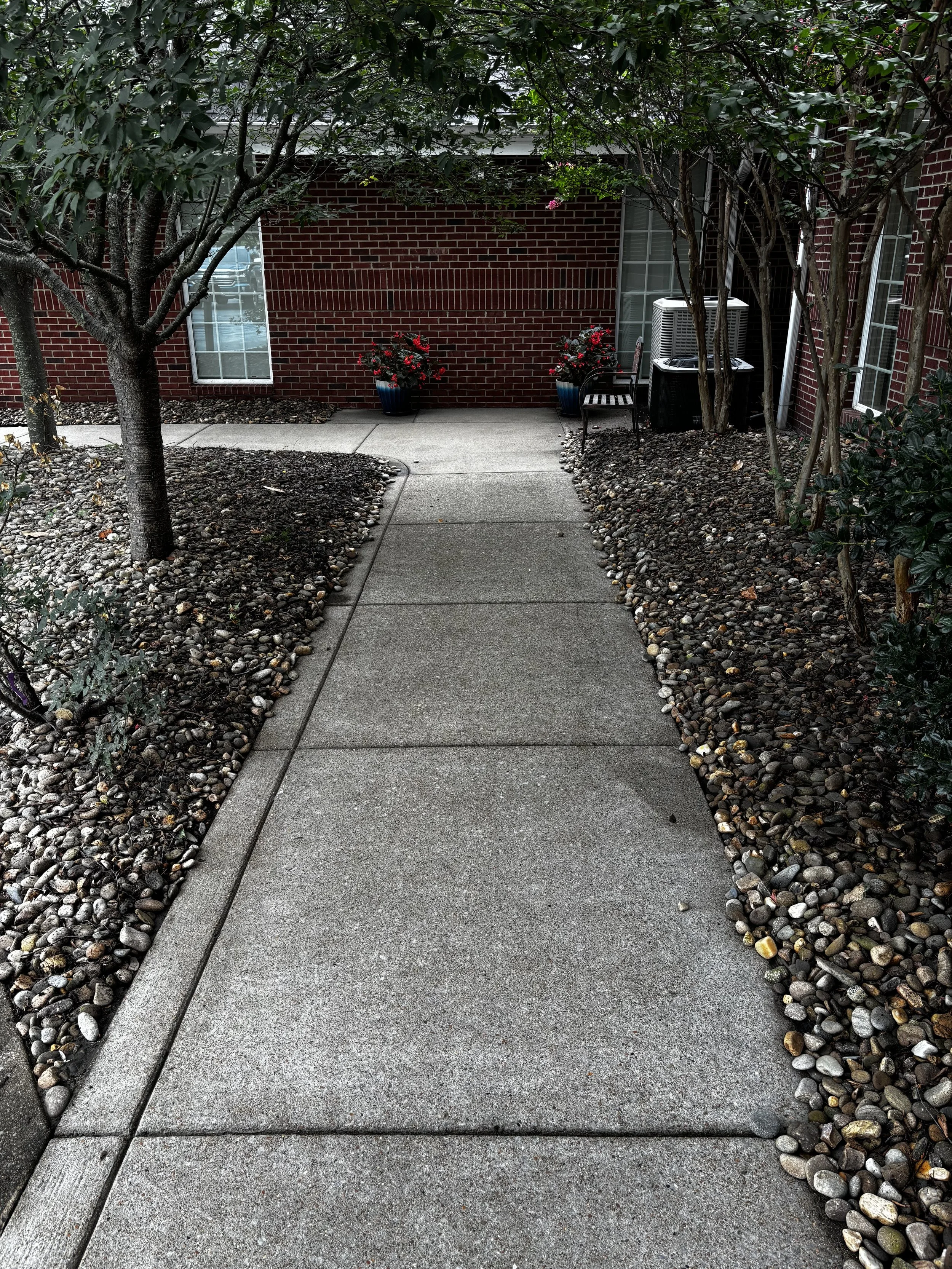 A concrete pathway leading to a red brick building with bushes and trees on both sides, two potted plants with red flowers on the back wall, a bench, and an air conditioning unit.