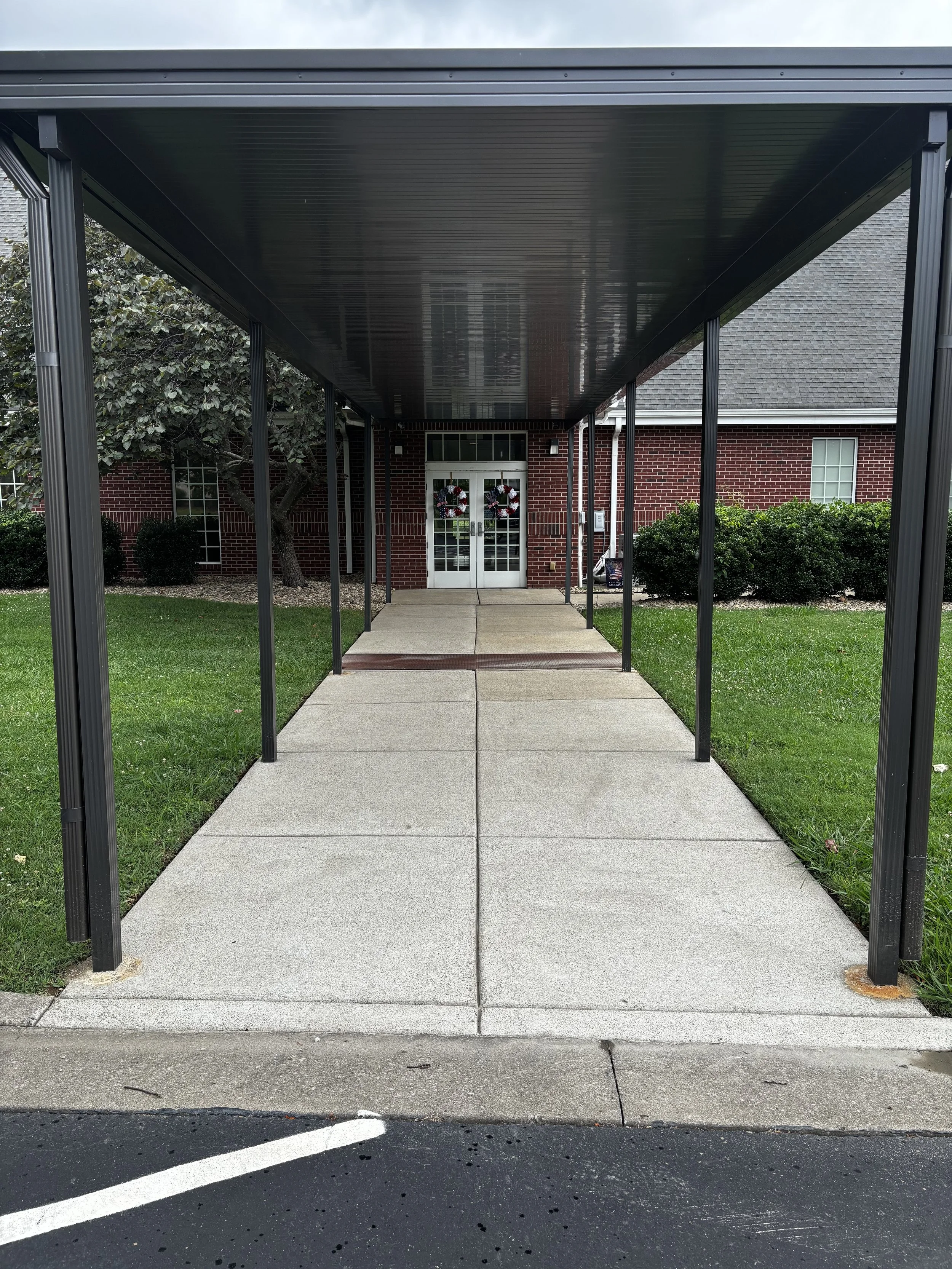 View of a sidewalk leading to a brick building with decorated Christmas wreaths on the door, covered by a metal awning supported by black metal posts. Green grass and bushes are on either side of the walkway, and the street with white parking lines i