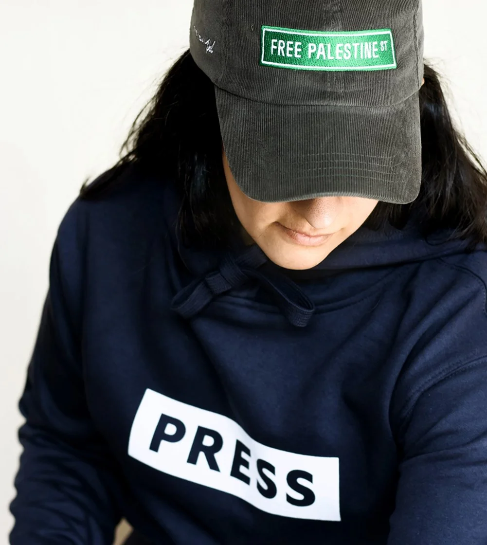 A person wearing a dark cap with a patch that reads 'Free Palestine,' and a navy hoodie with 'Press' written on it.