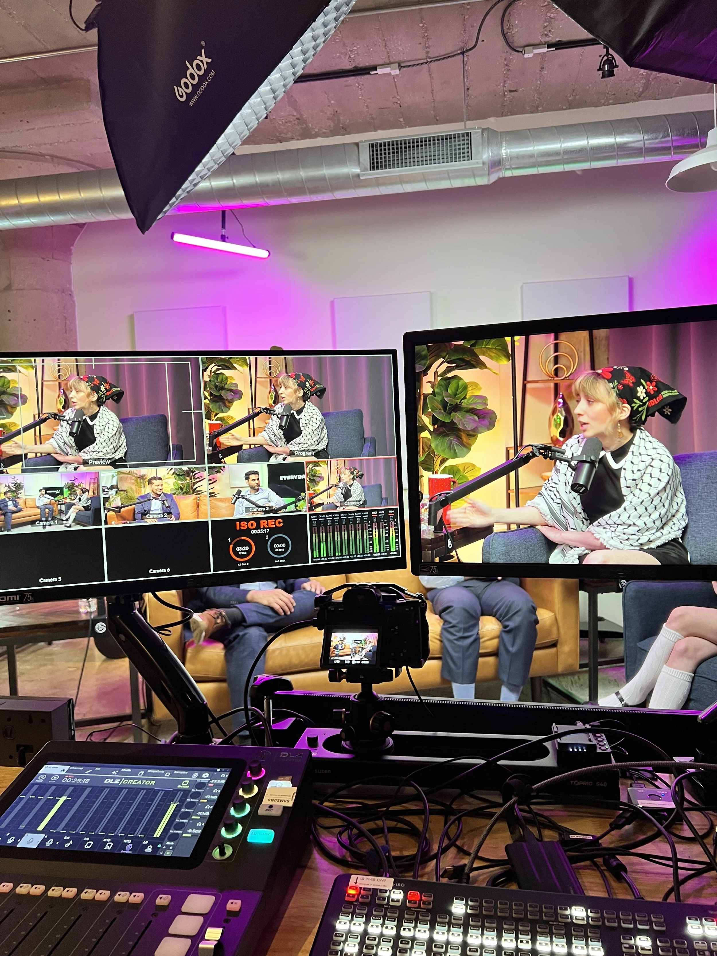 Recording studio or podcast set with monitors showing a woman in a headscarf speaking into a microphone, plants, and bright pink and purple lighting.