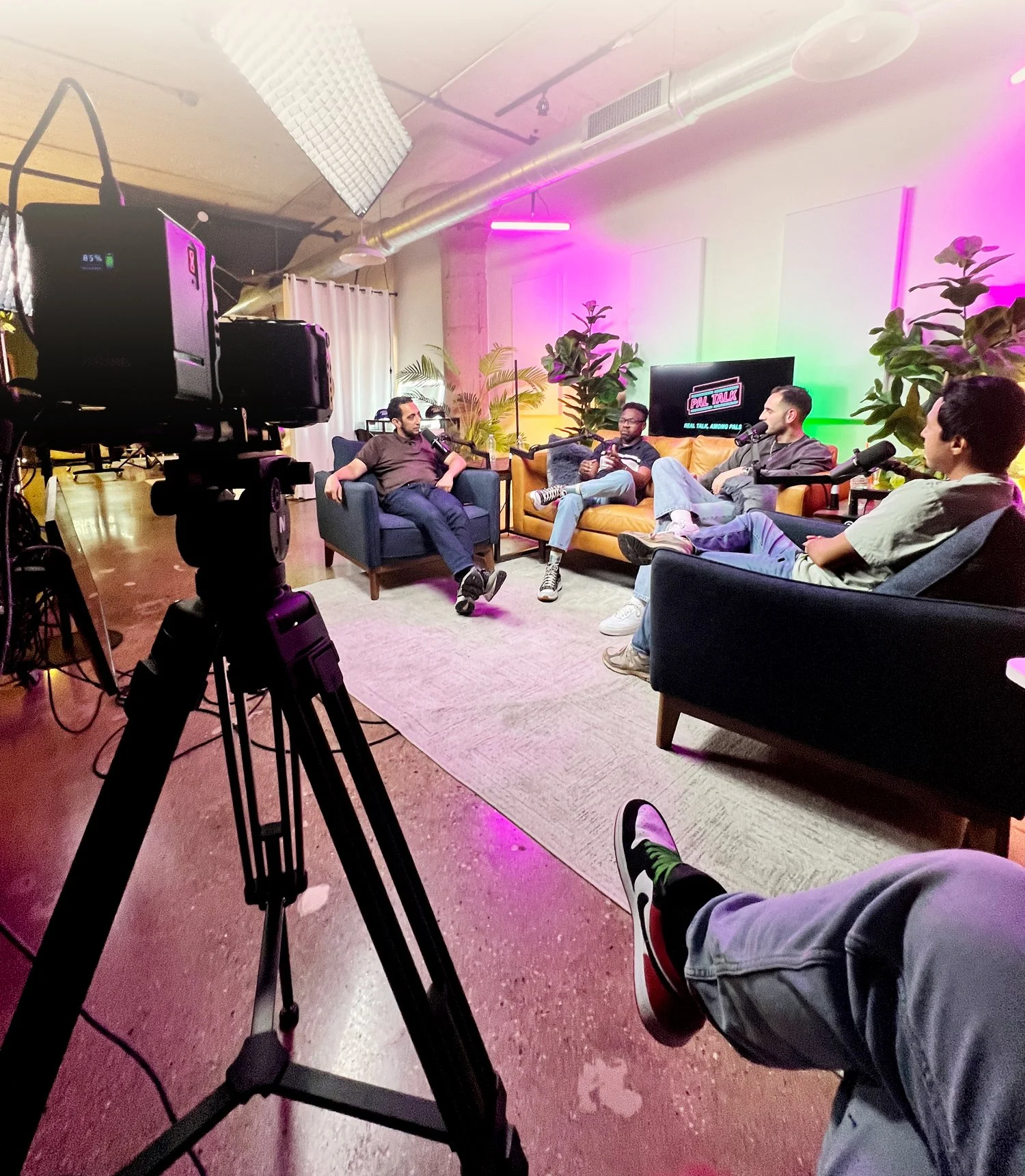Four people seated on couches in a recording studio or podcast room with colorful lighting, microphones, and a large camera filming them.