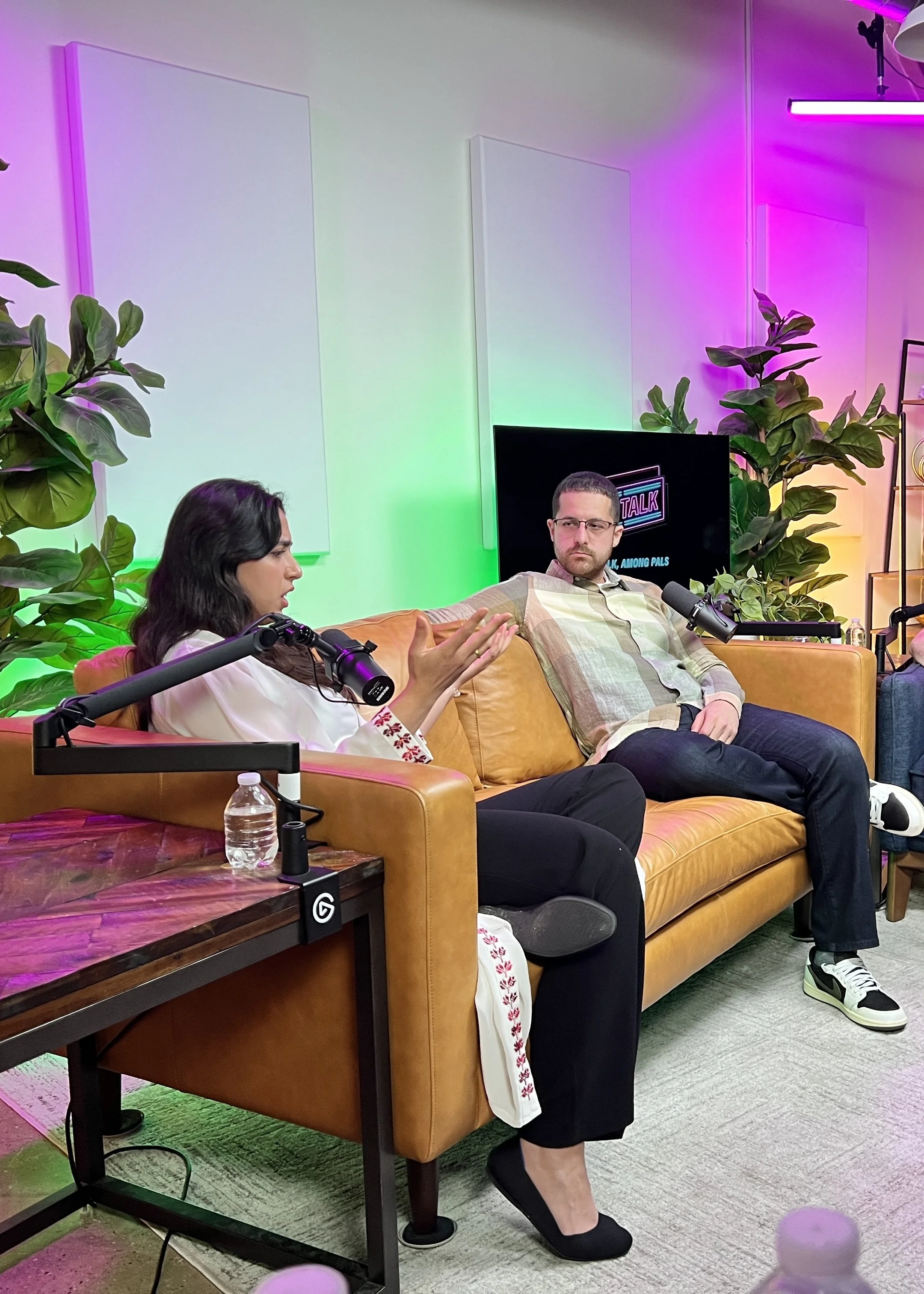 Two people sitting on a mustard-colored couch in a colorful, modern studio or podcast set. The woman on the left has long dark hair and is speaking into a microphone, gesturing with her hands. The man on the right has short dark hair, glasses, and is wearing a plaid shirt, listening attentively. The background features green, pink, and purple lighting, plants, and a TV screen with a logo that says 'TALK' and some text.