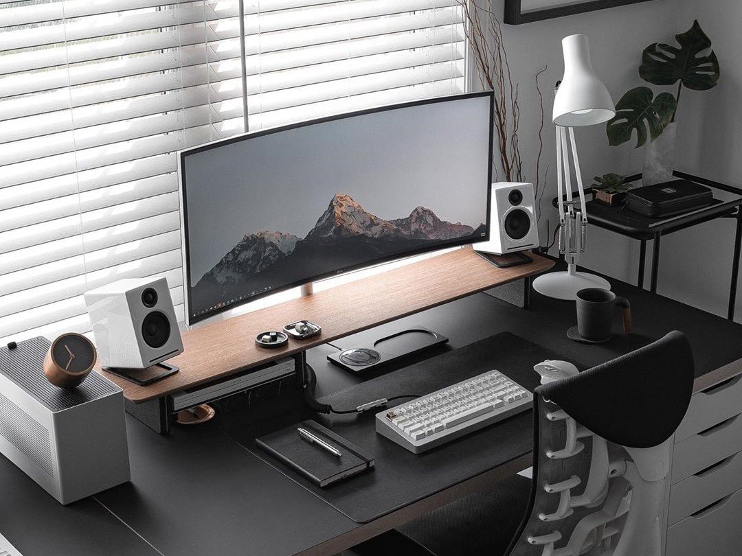 A modern computer workspace with a large curved monitor on a wooden stand, white speakers, a keyboard, mouse, and desk accessories on a black desk. There is a white lamp, a mug, a speaker, and potted plants in the background, with white blinds covering a window.