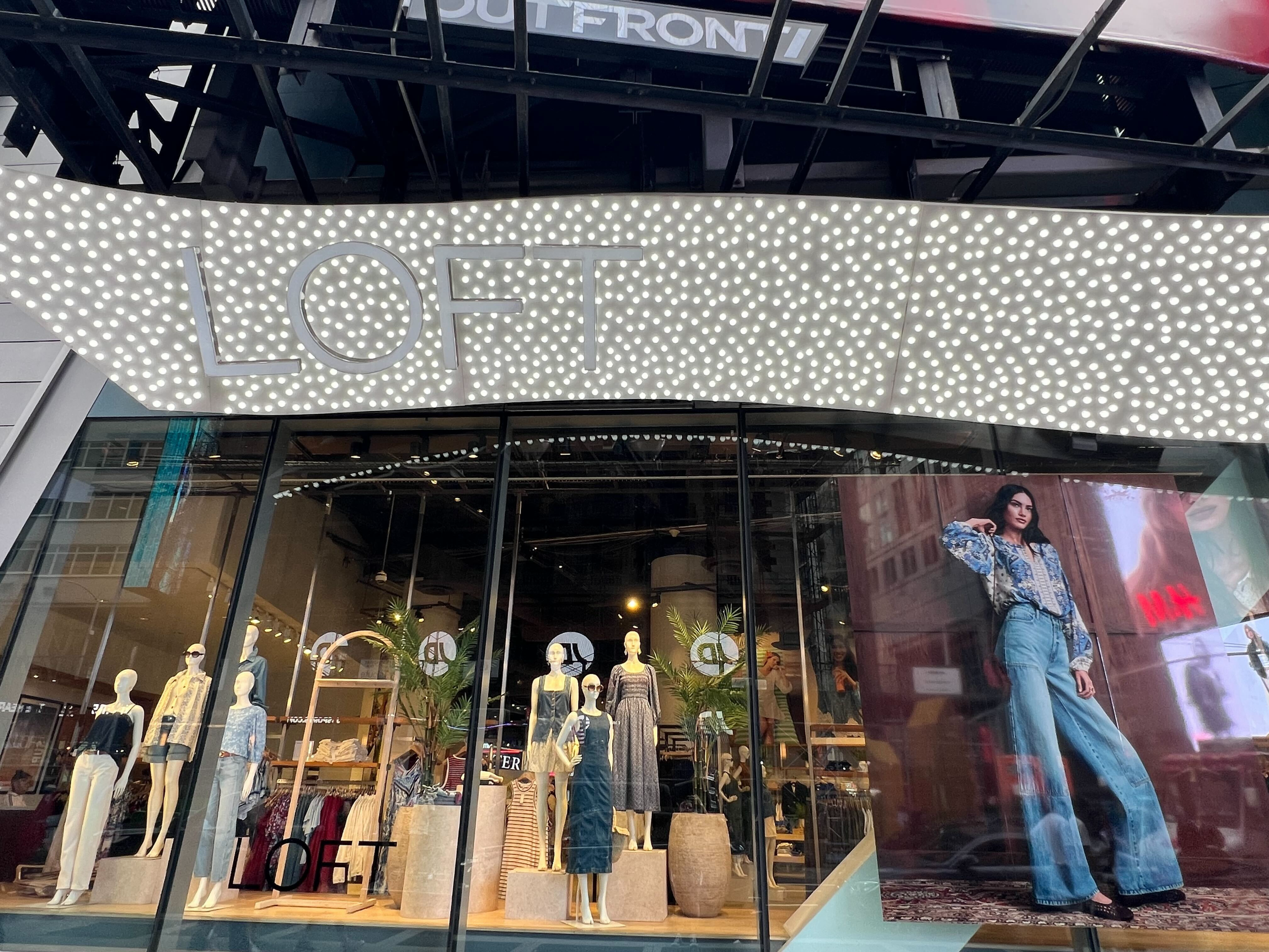 Clothing store window display with mannequins dressed in casual wear, bright white lights overhead, and a large signs that reads 'LOFT' outside a shopping mall.