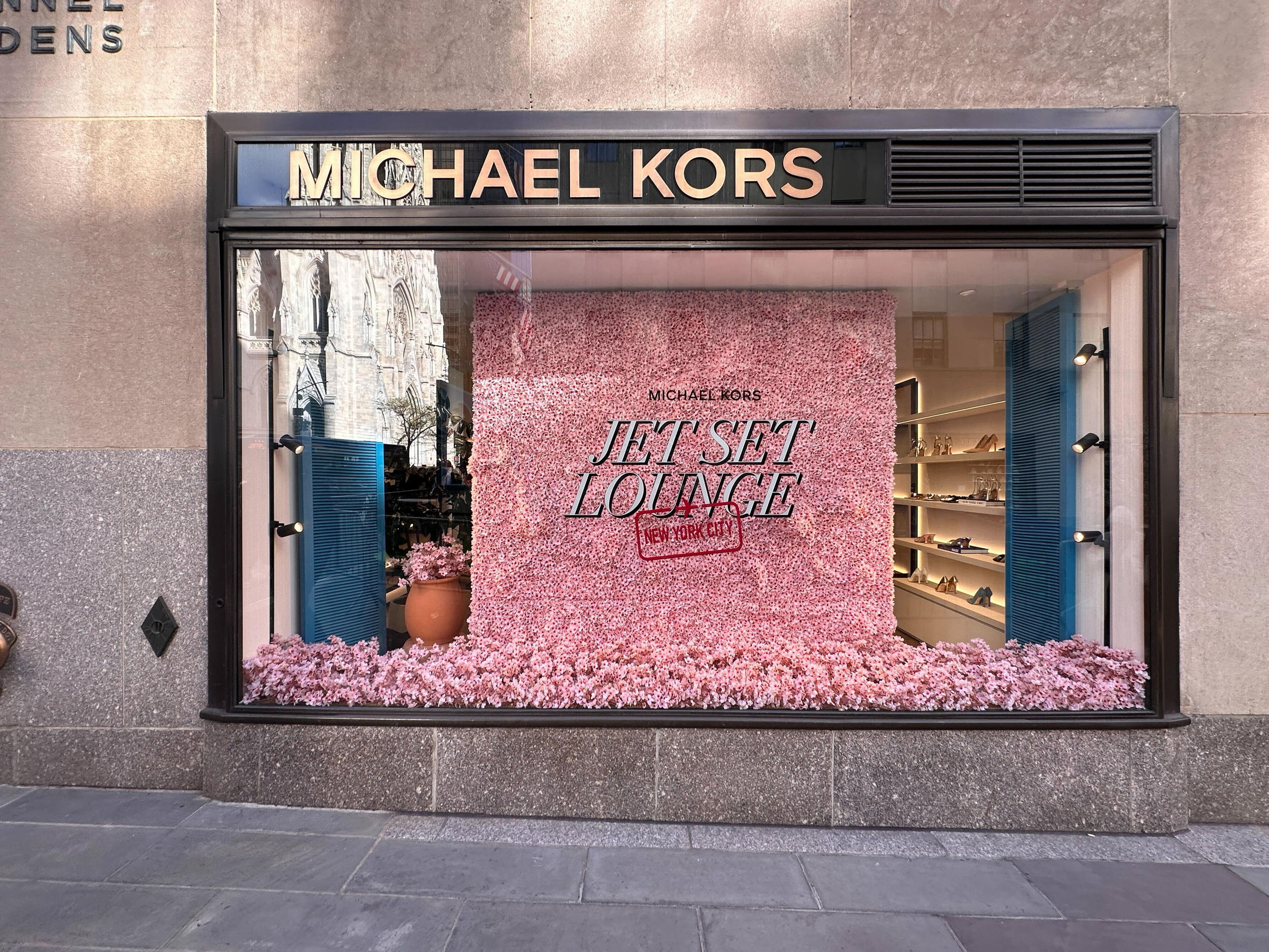 Storefront window display with the text 'Jet Set Lounge' surrounded by pink flowers and a 'New York City' stamp, featuring shoes and accessories inside.
