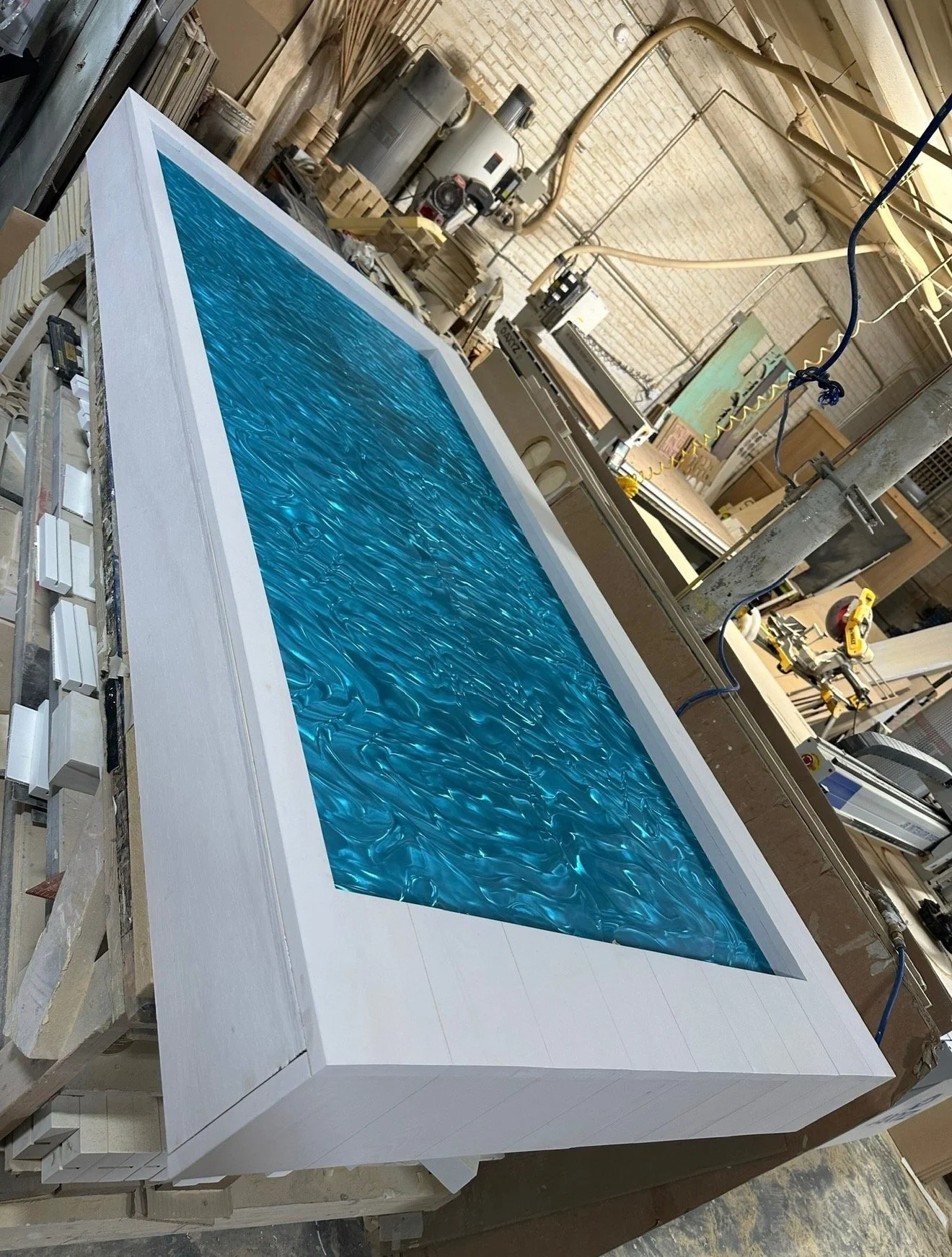 A large rectangular indoor pool with a textured blue water pattern, surrounded by a white frame, situated in a woodworking or carpentry workshop filled with tools, machinery, and wood materials.