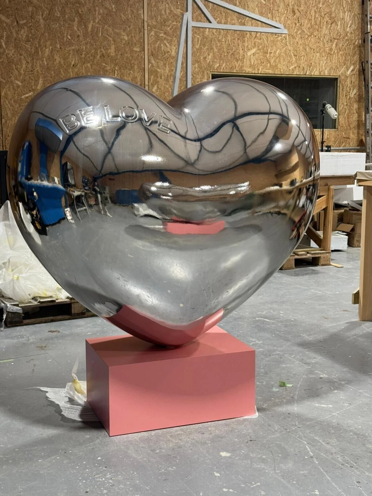 A shiny, reflective, silver heart-shaped sculpture with the word 'BELIEVE' engraved on it, displayed on a pink pedestal in a warehouse or studio with wooden and metal structures and equipment.