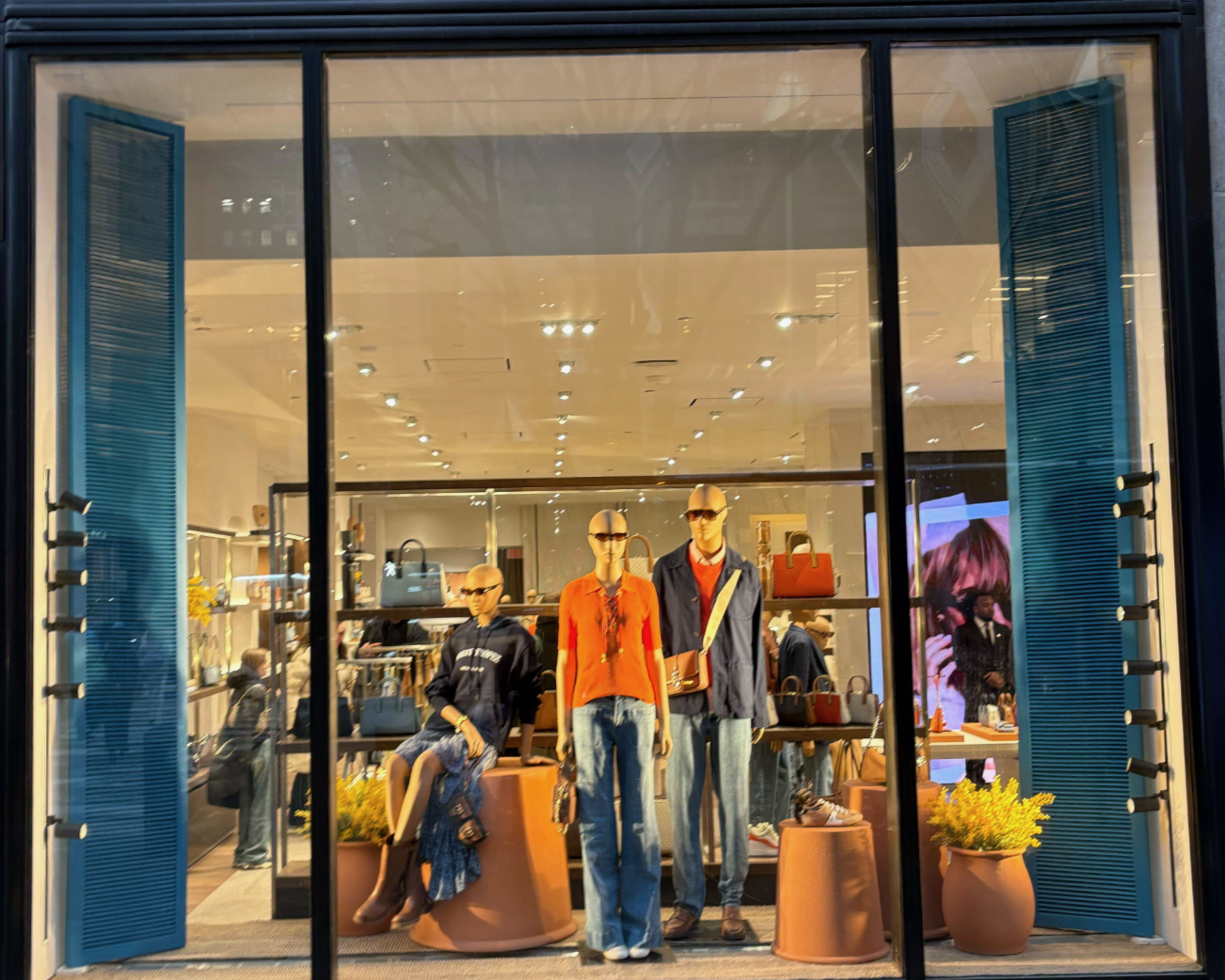 View of a clothing store window display featuring three mannequins dressed in casual outfits, with shoes and handbags on display, and potted plants in front of the window.