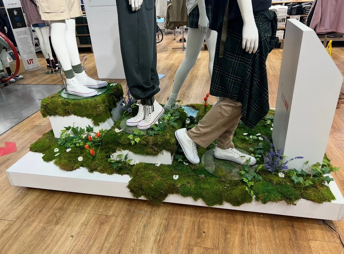 Mannequins dressed in casual shoes on a mossy, nature-themed display with flowers and greenery inside a retail store.