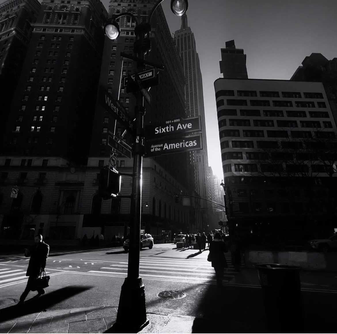 A city street scene in black and white showing tall buildings, street signs for Sixth Avenue and the Americas, and pedestrians crossing the street in sunlight.
