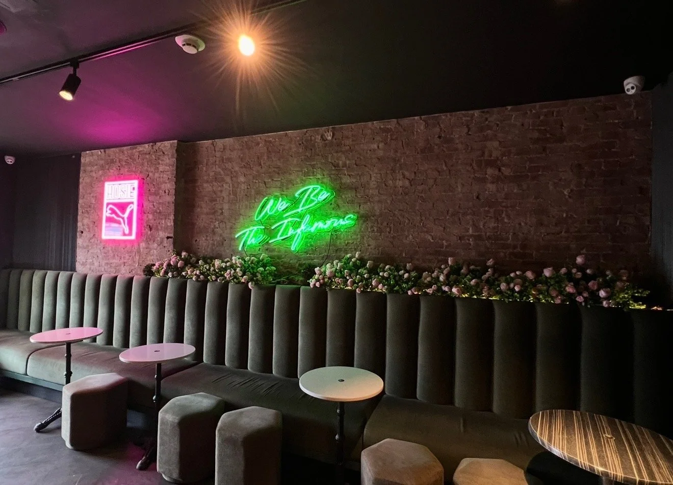 Interior of a lounge or bar with brick wall, featuring neon signs reading 'House' with a cat logo and 'We Are The Infamous.' There is a long upholstered bench with small tables and stools, and a floral arrangement along the wall.