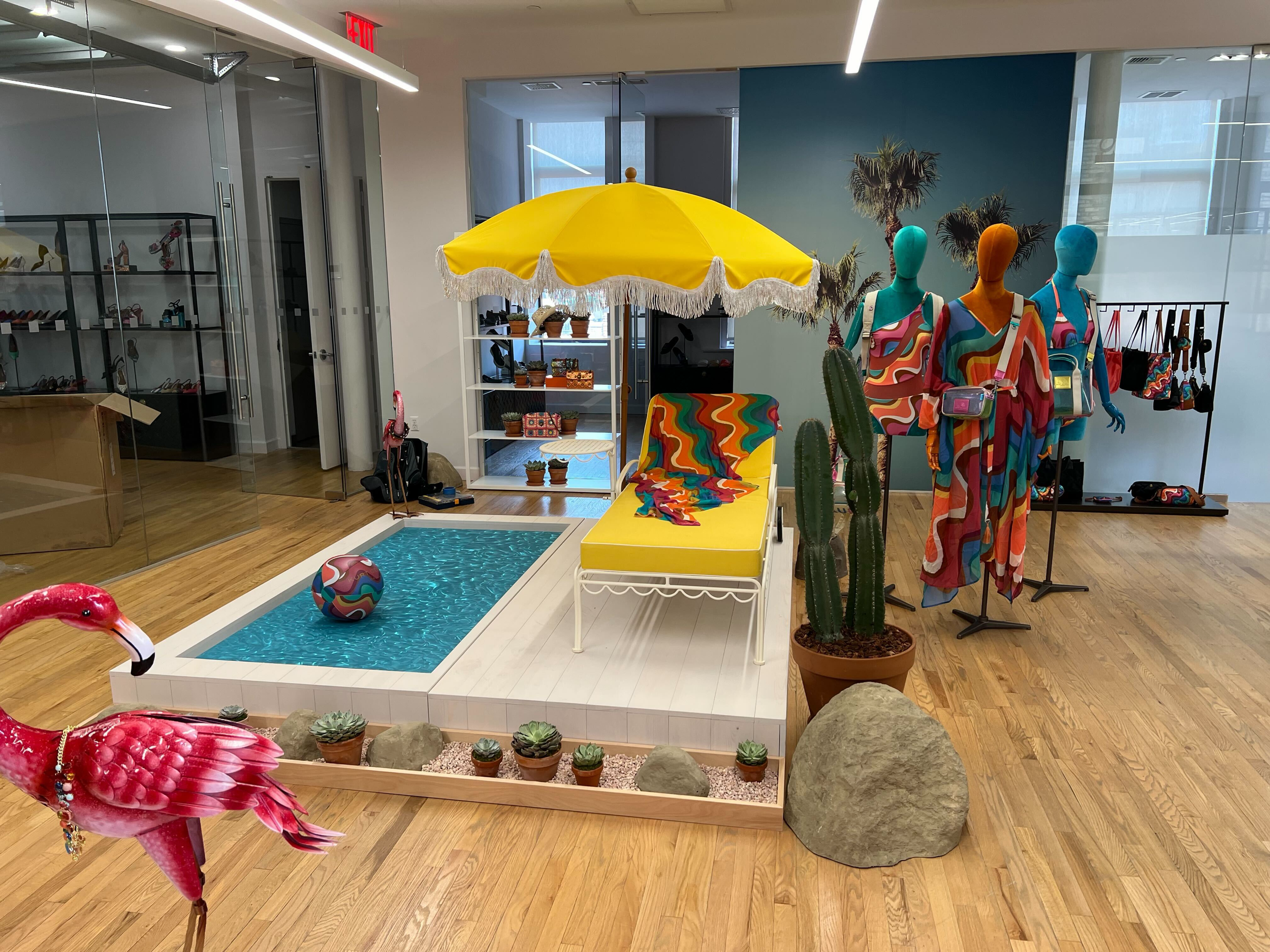 Display of colorful mannequins wearing vibrant, psychedelic-patterned clothing inside a retail shop. A yellow umbrella with tassels is positioned behind them, and a cactus plant is nearby. The store has a modern, well-lit interior with a wood floor a