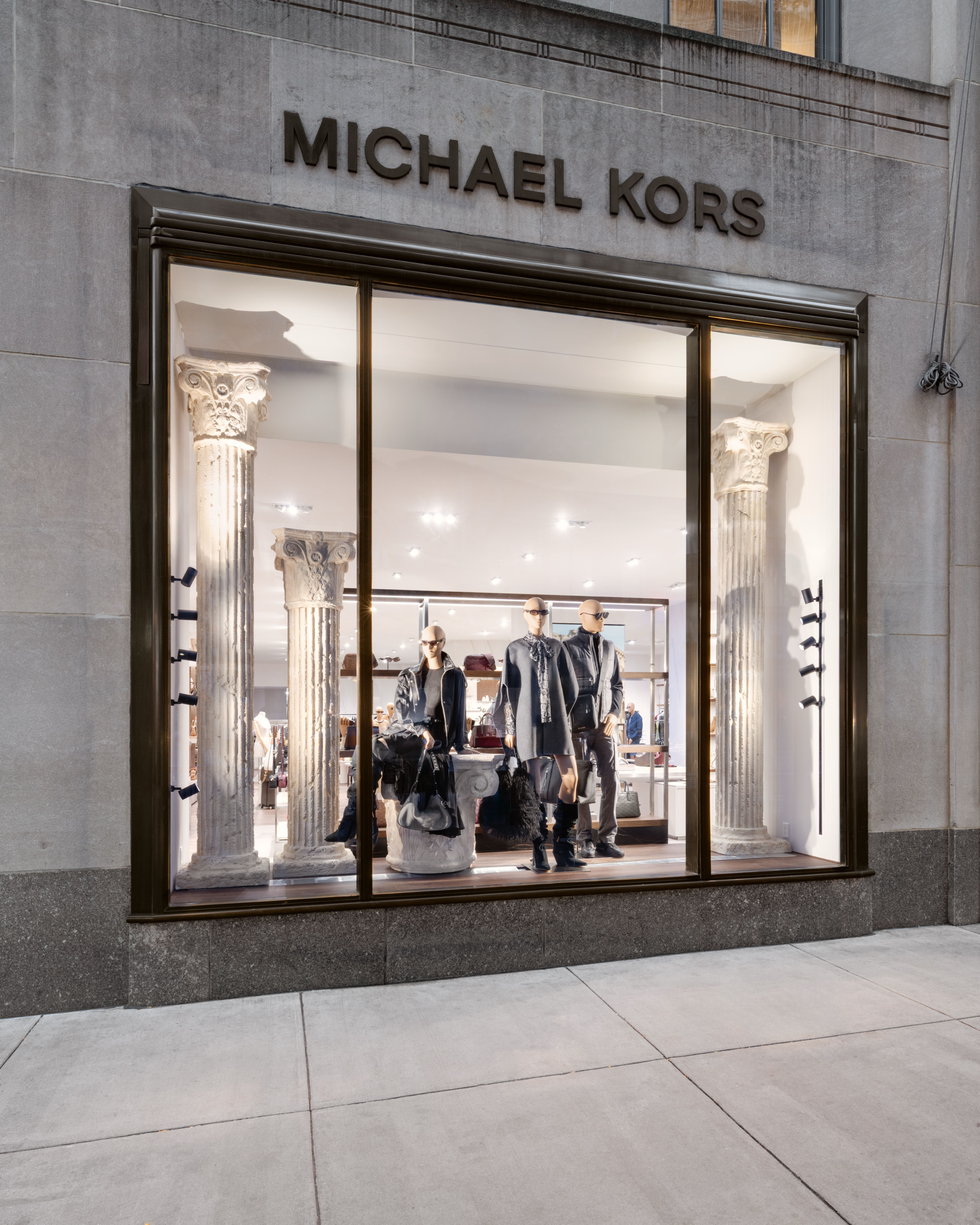 Shop window display at Michael Kors with mannequins dressed in clothing and accessories, framed by several classical columns within a modern storefront.