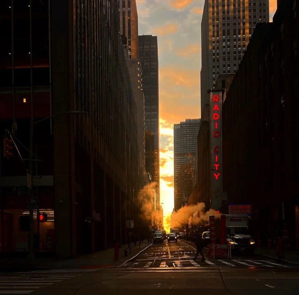 Urban street scene during sunset with tall buildings on both sides, steam rising from street vents, and a neon sign reading 'Radio City' on the right. Vehicles and pedestrians are visible on the street.