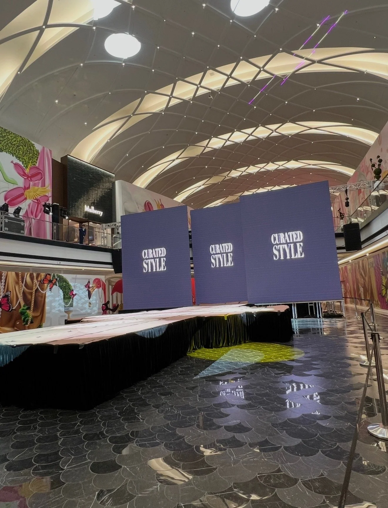 Interior of a shopping mall with a stage setup and large digital screens displaying the words 'Curated Style'. The ceiling has an illuminated, arched design, and some decorative wall art with floral and butterfly motifs is visible in the background.