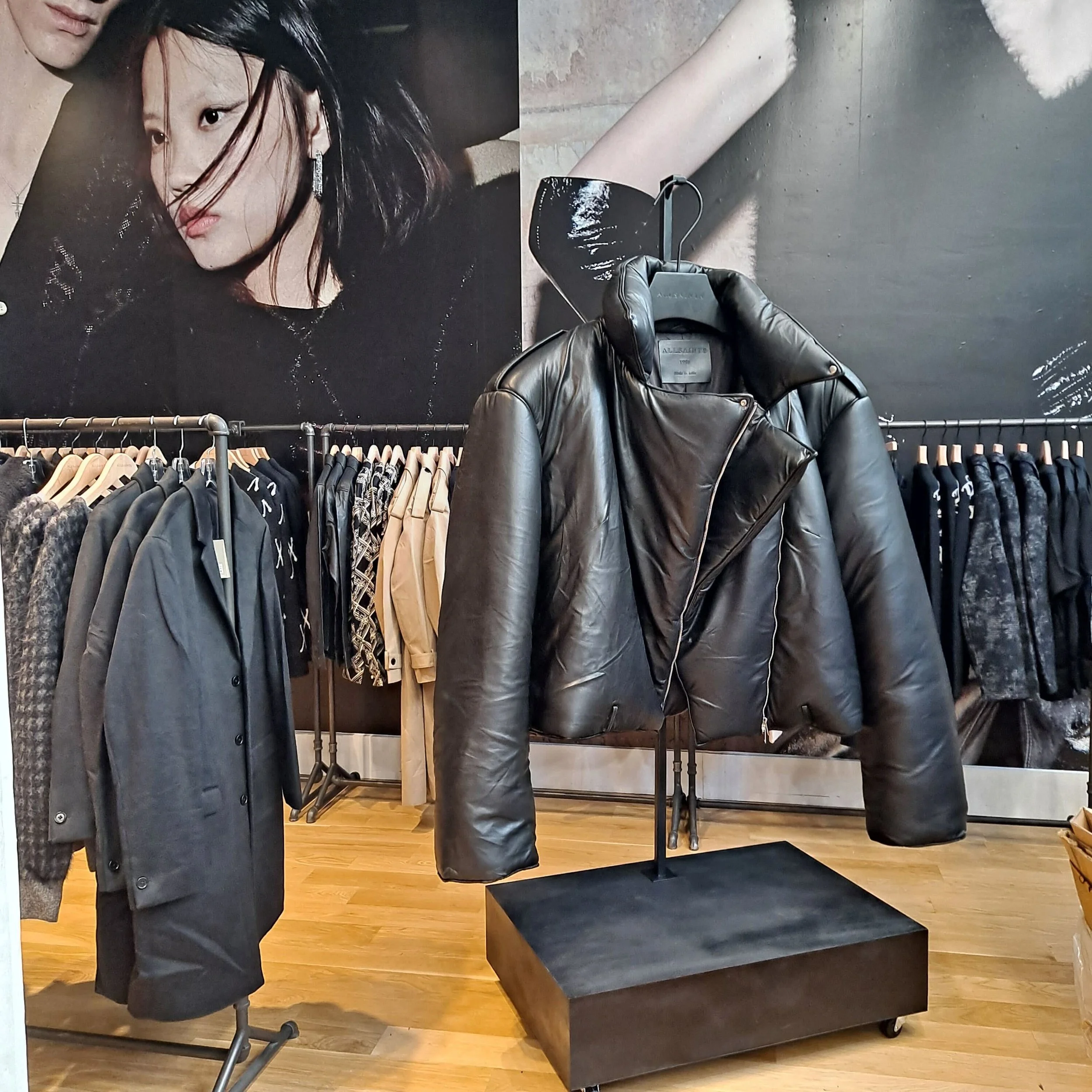 A large fashion advertisement poster of a woman with black hair, wearing black earrings, on a wall in a clothing store. The store has a display rack with various black and beige clothing items, a black leather jacket on a hanger, and a wooden floor.