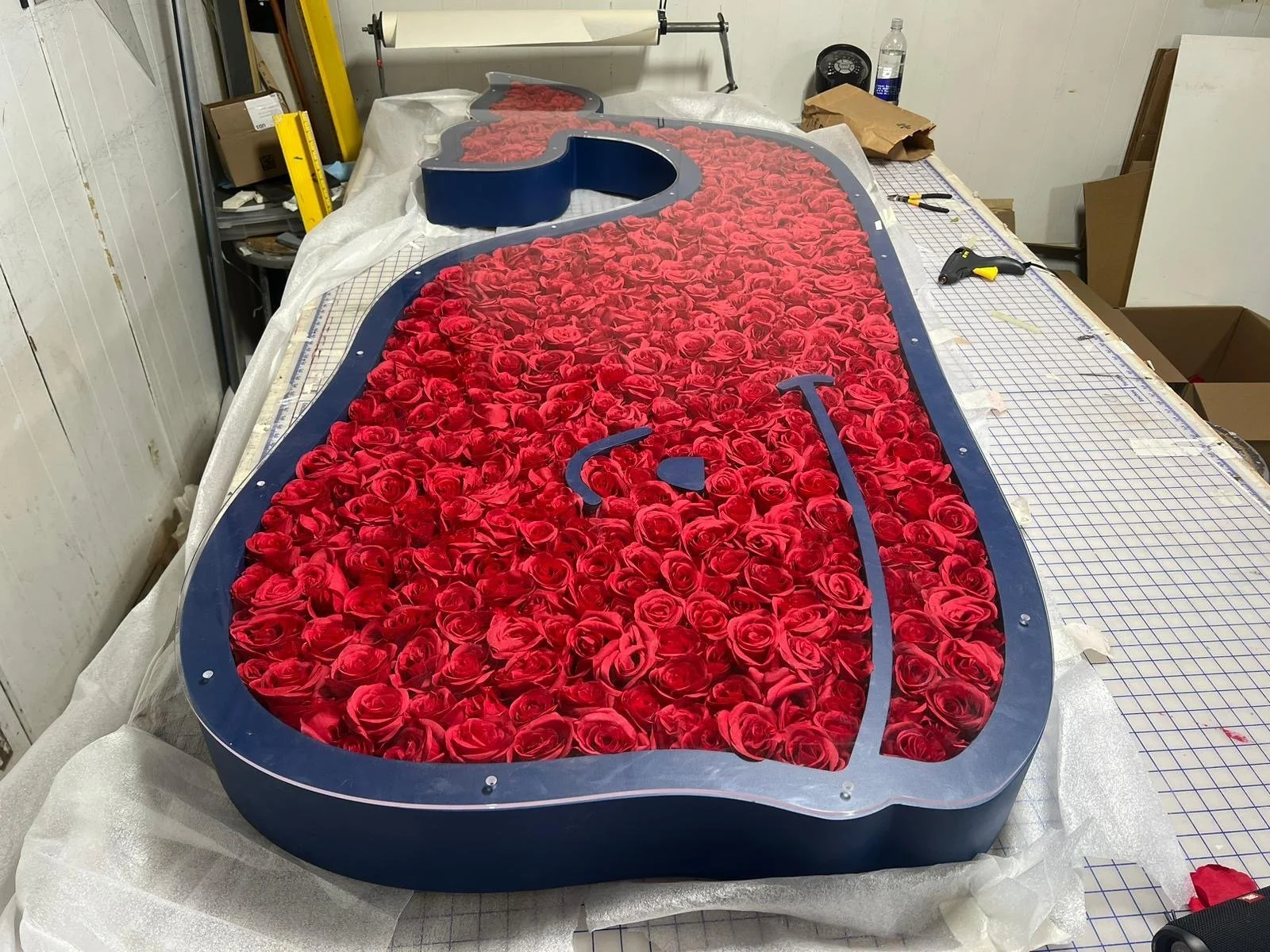 A large decorative sign filled with red roses, with a blue border and metallic backing, being assembled on a worktable.