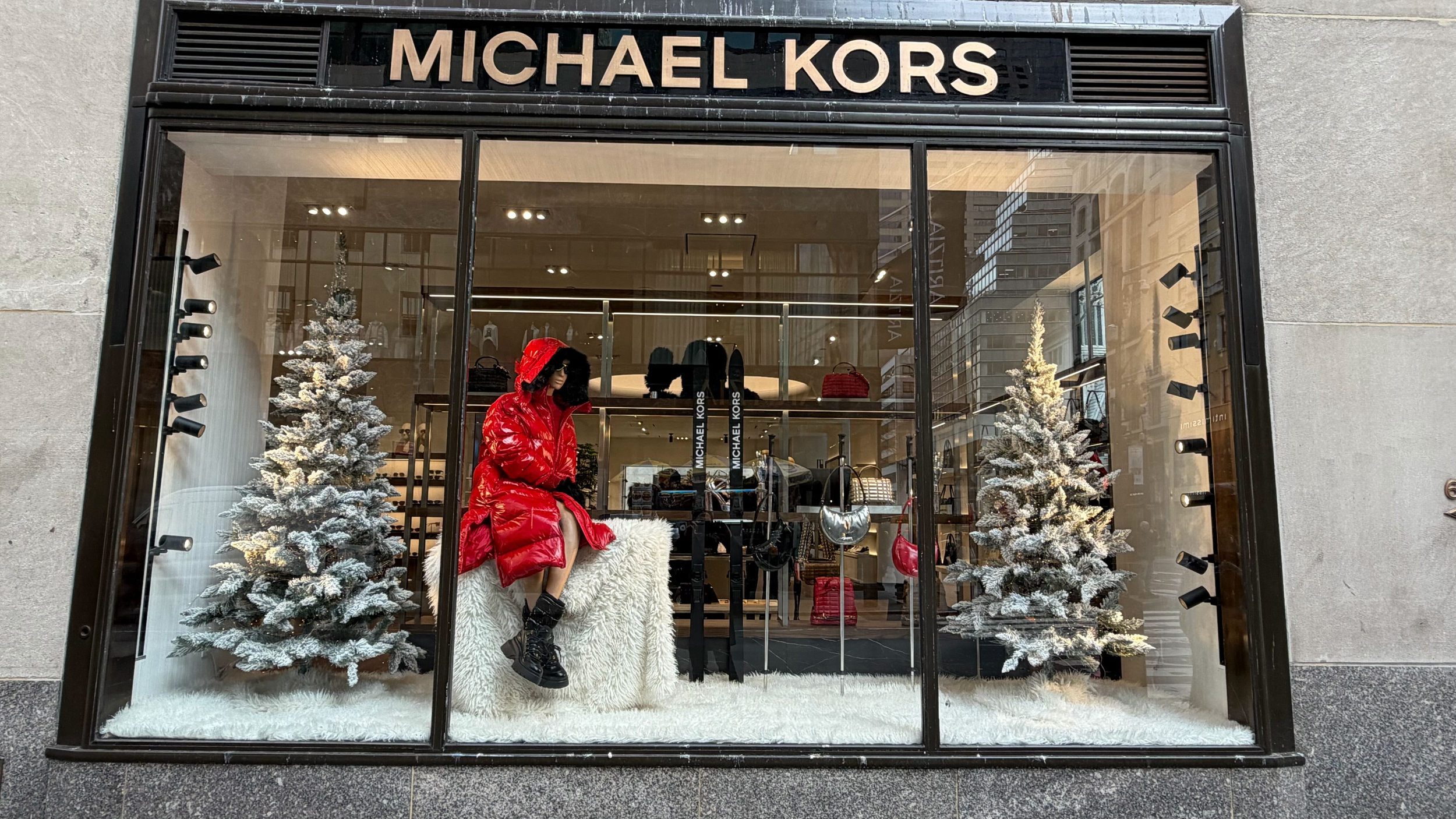 A retail store window display for Michael Kors featuring a mannequin dressed in a red puffer coat, black boots, and sunglasses, with two snow-dusted artificial Christmas trees and handbags inside the store.