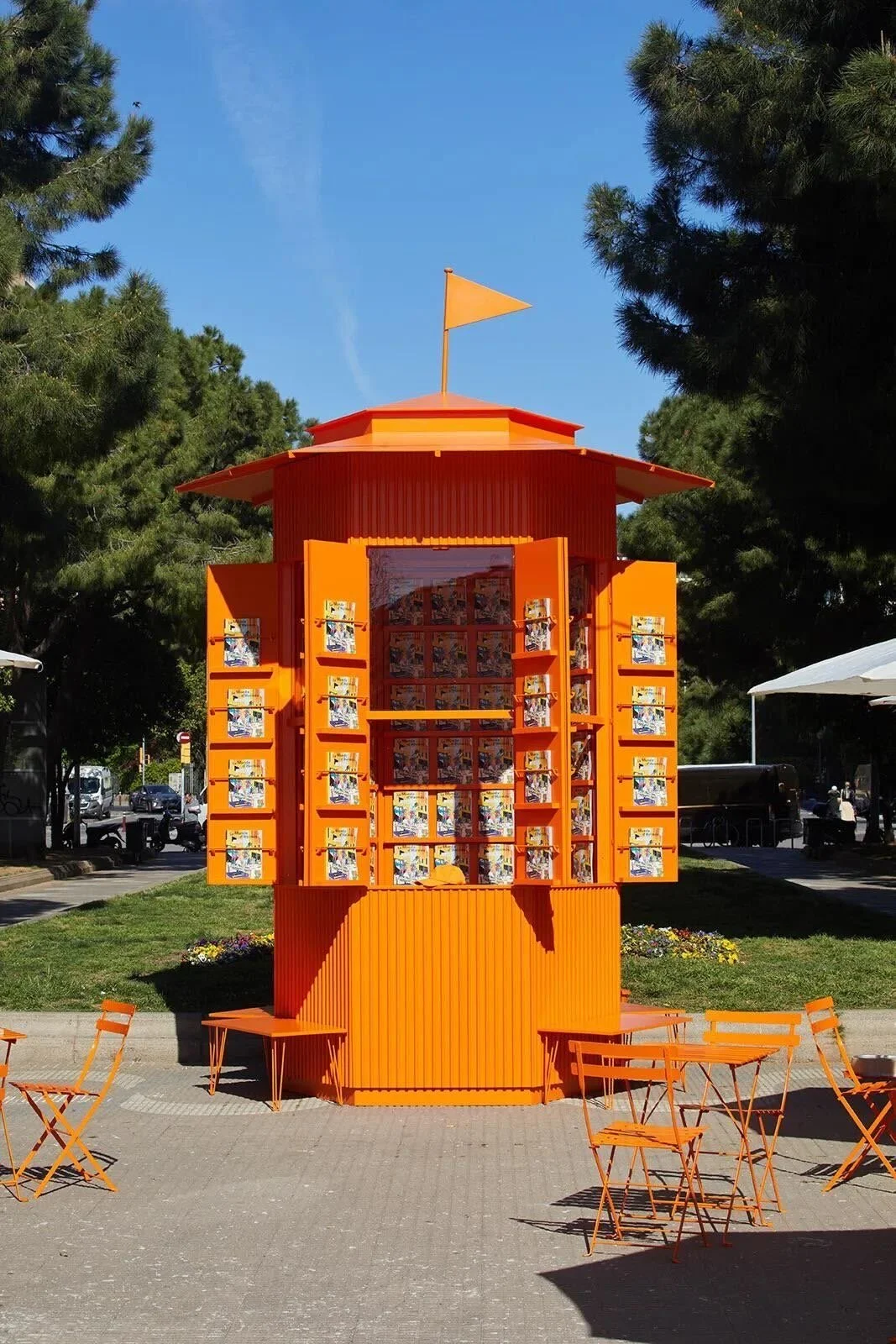 Orange kiosk with open shutters displaying magazines, surrounded by orange chairs and tables, set outdoors on a sunny day with trees and park in the background.