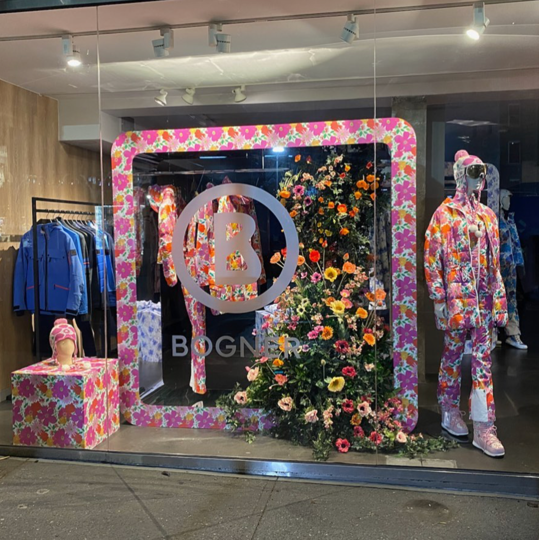 Store window display featuring mannequins dressed in pink floral pajamas, surrounded by a large floral tree and decorated with a pink floral frame and table.