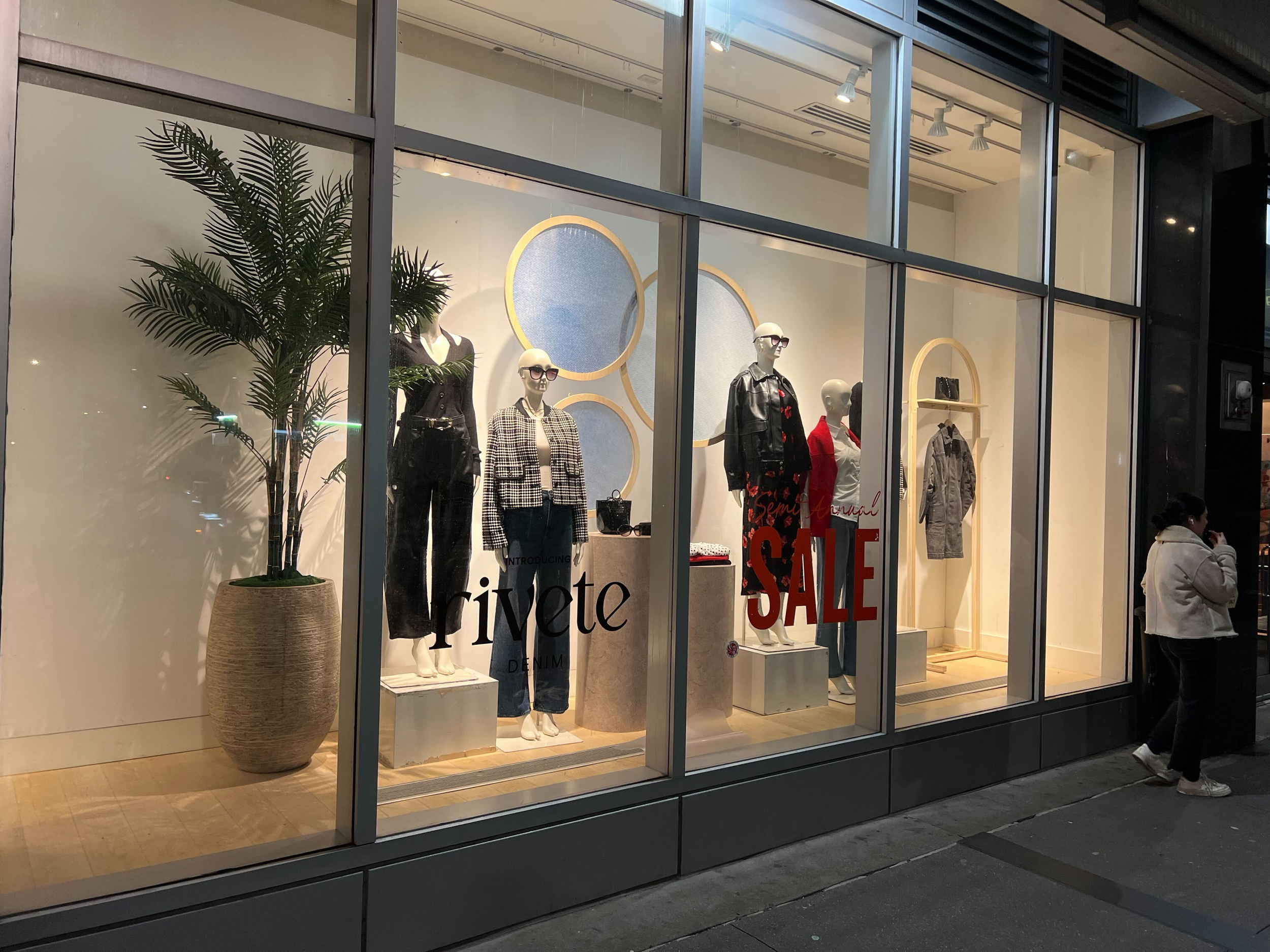 Store window display with mannequins dressed in fashionable clothing, large decorative circles on the wall, a potted plant on the left, and a sale sign with red letters.
