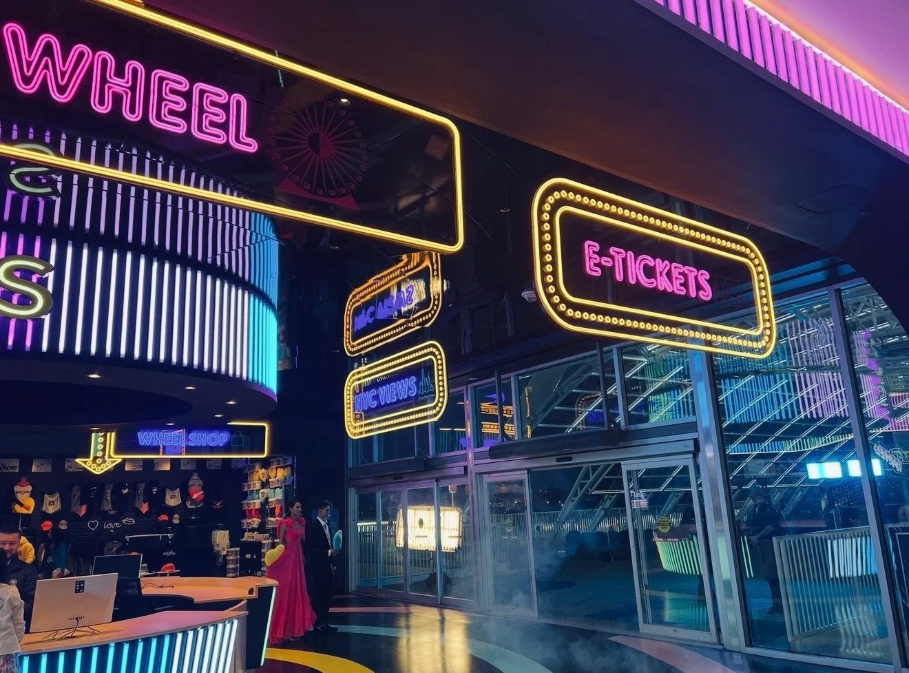 Neon signs indicating ticket and wheel shop entrances at an amusement park, with people near the entrances and a colorful roller coaster in the background.