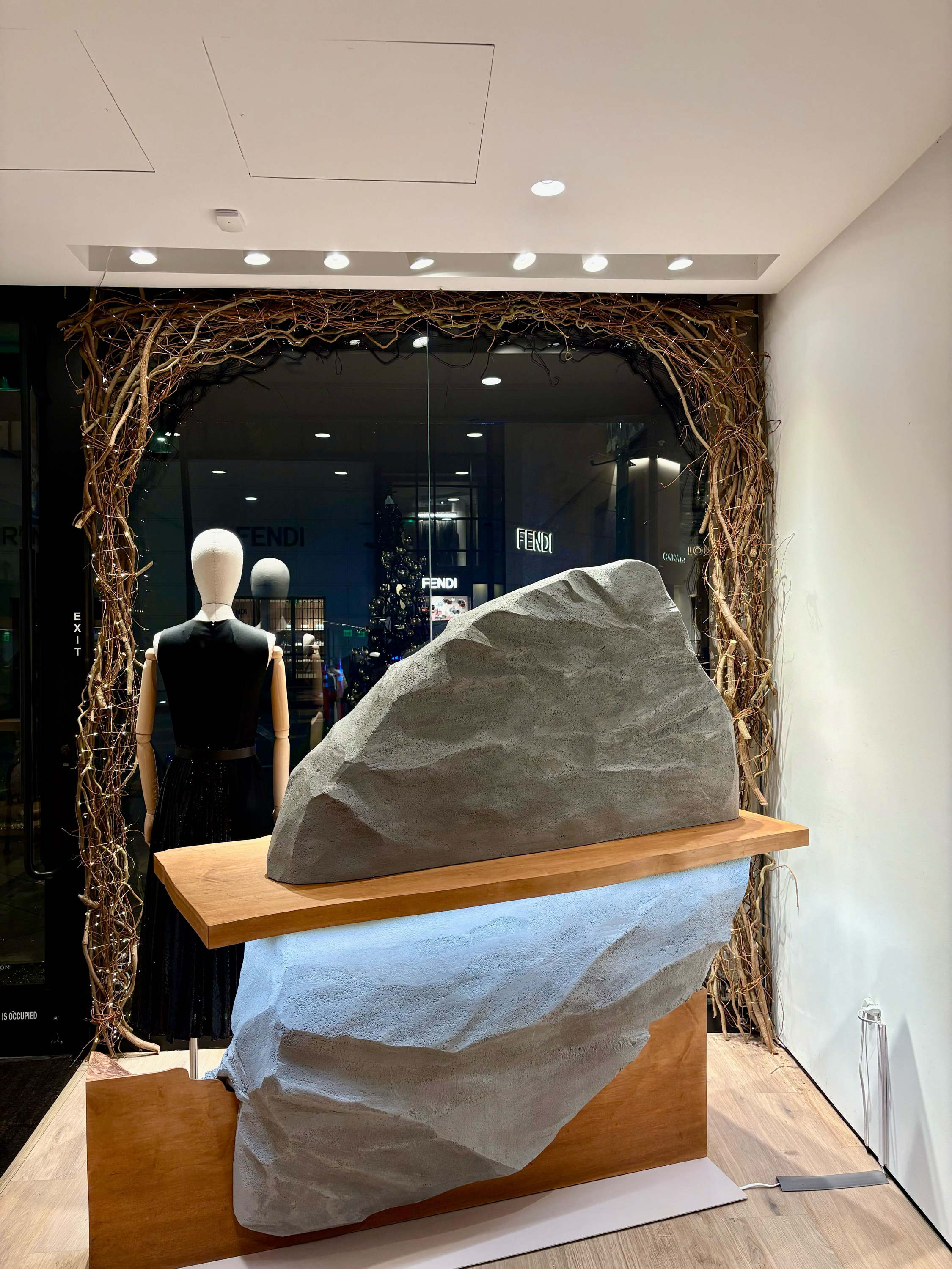 A display with a large, jagged, gray stone sculpture on a wooden base and a wooden shelf. Behind the sculpture is a woman mannequin dressed in a black sleeveless dress. The display is framed by a border made of twisted, leafless branches. In the back